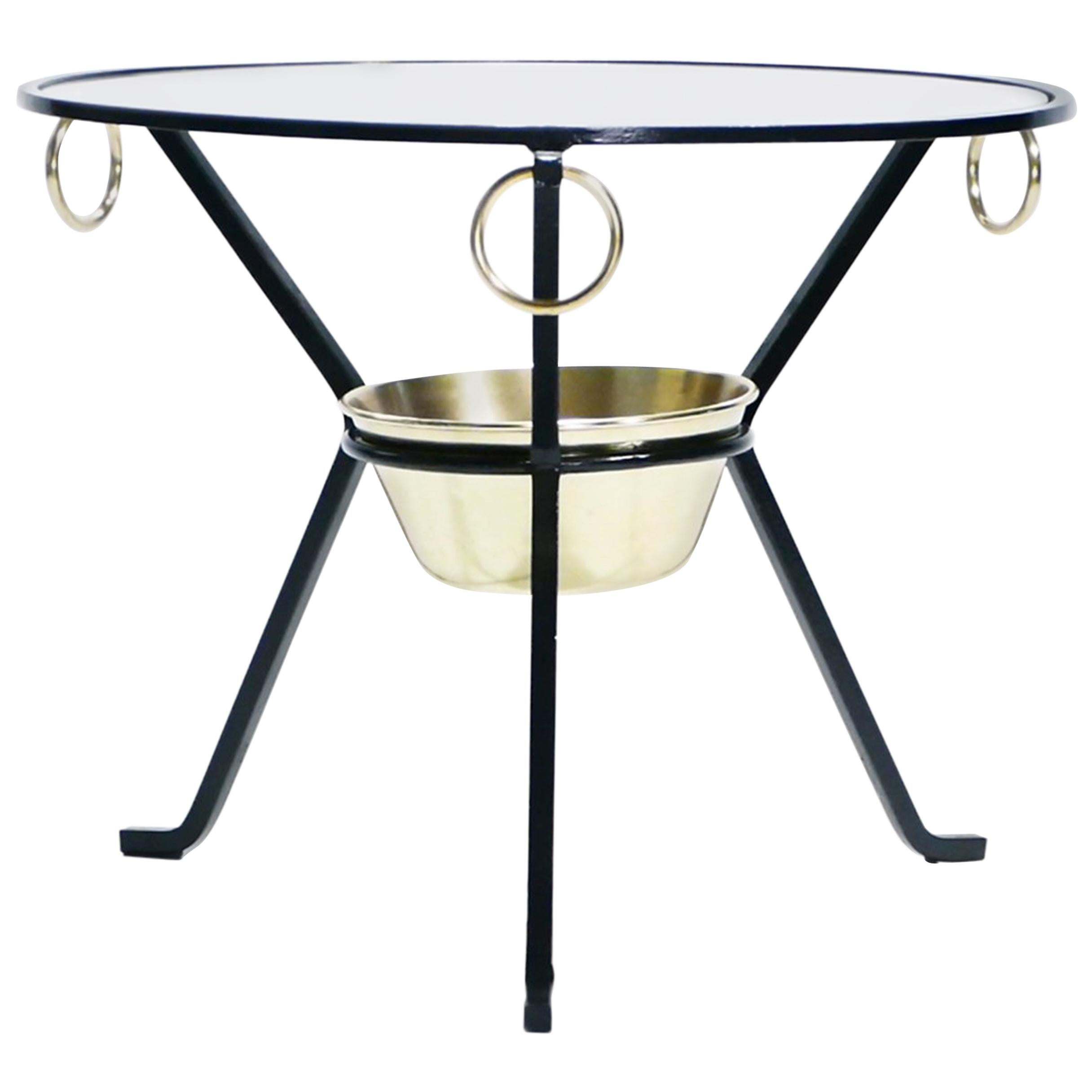 A sophisticated blend of Mid-Century Modern and neoclassical design, this 1950s side table or gueridon by Jacques Adnet features bold brass details, matte black metal feet, and a transparent glass top. Triple legs cross gracefully to support the
