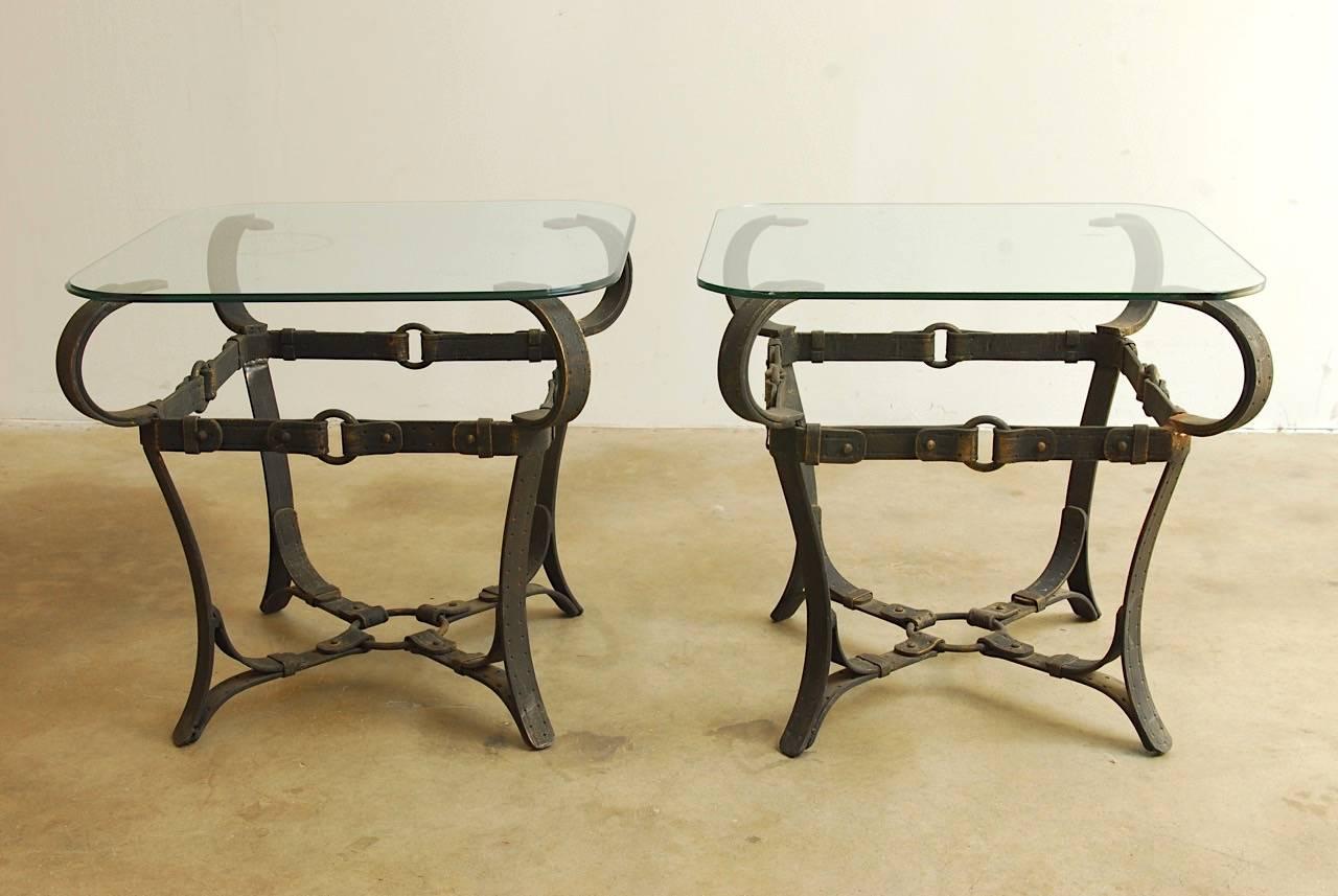 Chic pair of end tables made in the manner of Adnet. Constructed from unique cast iron faux leather equestrian straps, rings, and buckles. Classic intricate details and rich patina with a nod to Hermes. Each has a square glass pane with rounded