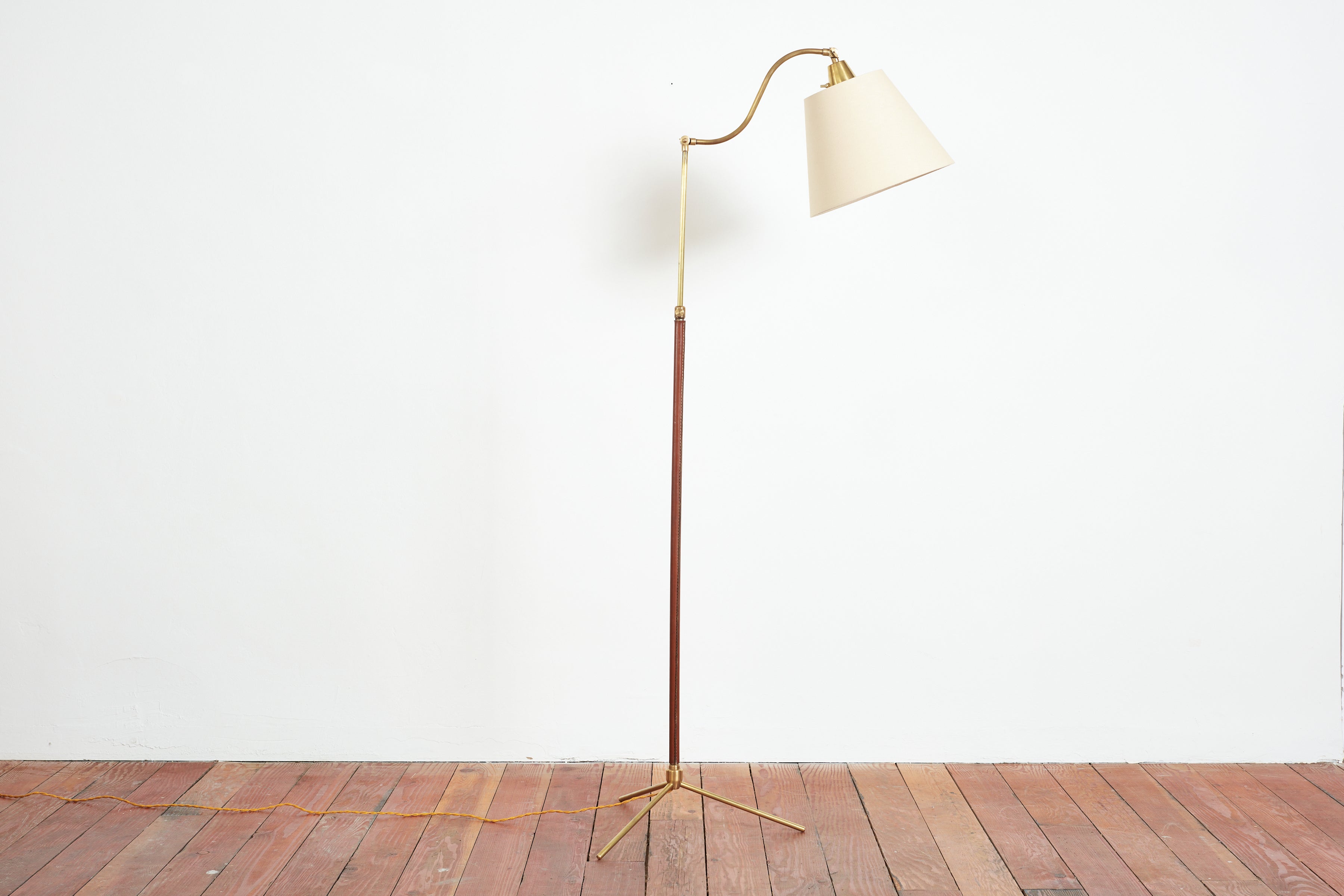 Handsome French floor lamp by Jacques Adnet with hand-stitched brown leather, contrast stitching, brass tripod base and new linen shade.

Unique arched and articulating neck that adjusts in height and gracefully angles downward. 

Height extends