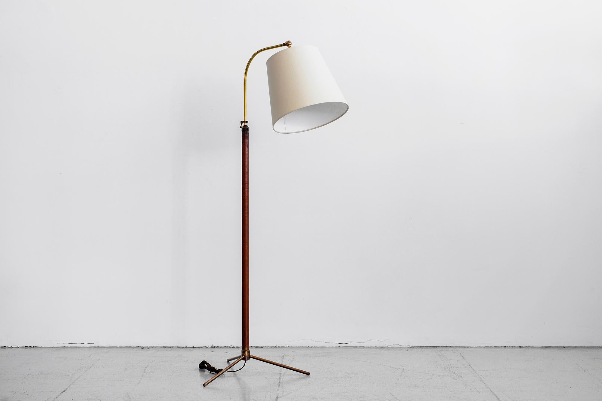 Handsome French floor lamp in the style of Jacques Adnet. Brass stem wrapped in saddle brown leather with contrast stitching and brass tripod base. Great age and patina. Shade pivots and brass neck is height adjustable. Newly re-wired with new shade.