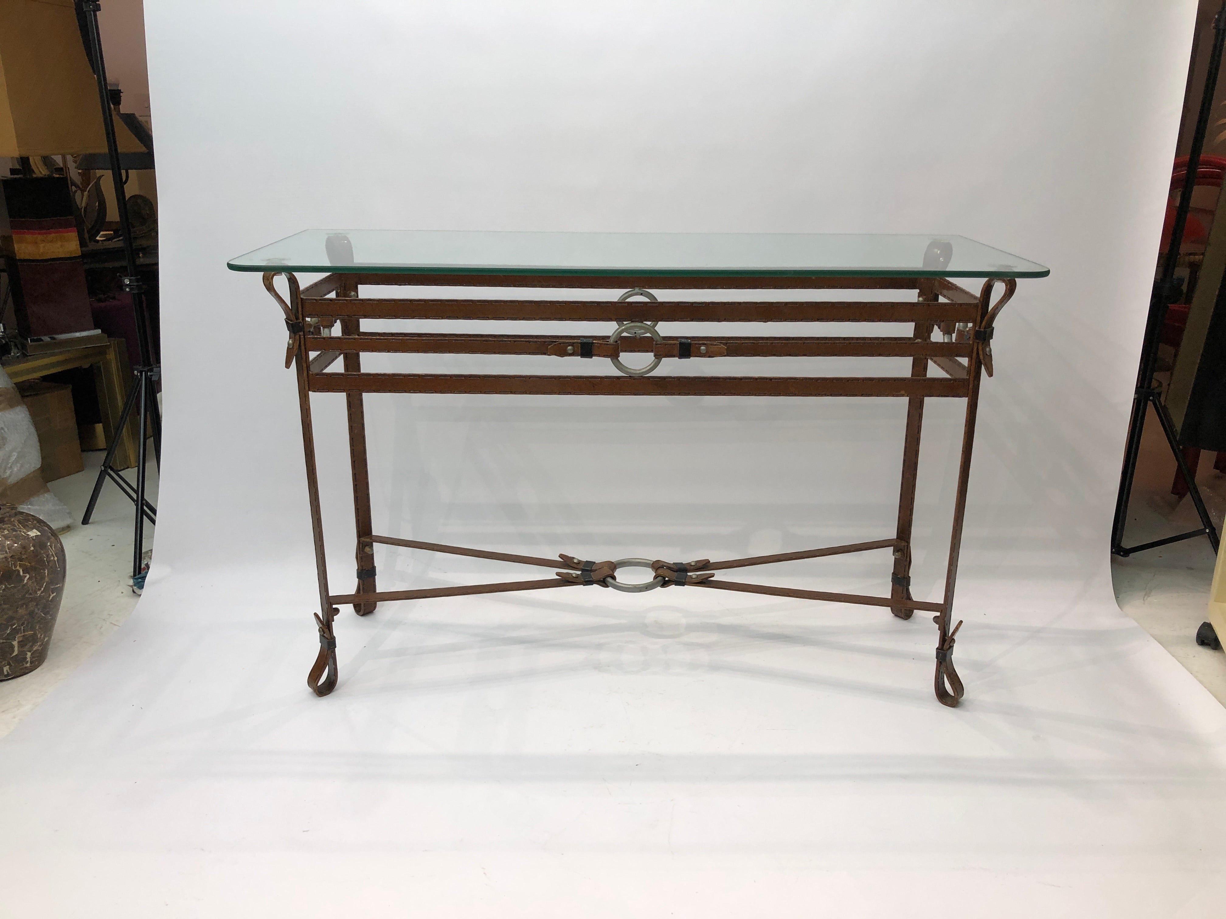 Iron frame console table inspired by Jacque Adnet leather and stitch designs.
This console table represents brown leather stitched straps that are joined by iron rings and they form a pleasing design that supports the glass top. 
The glass top has