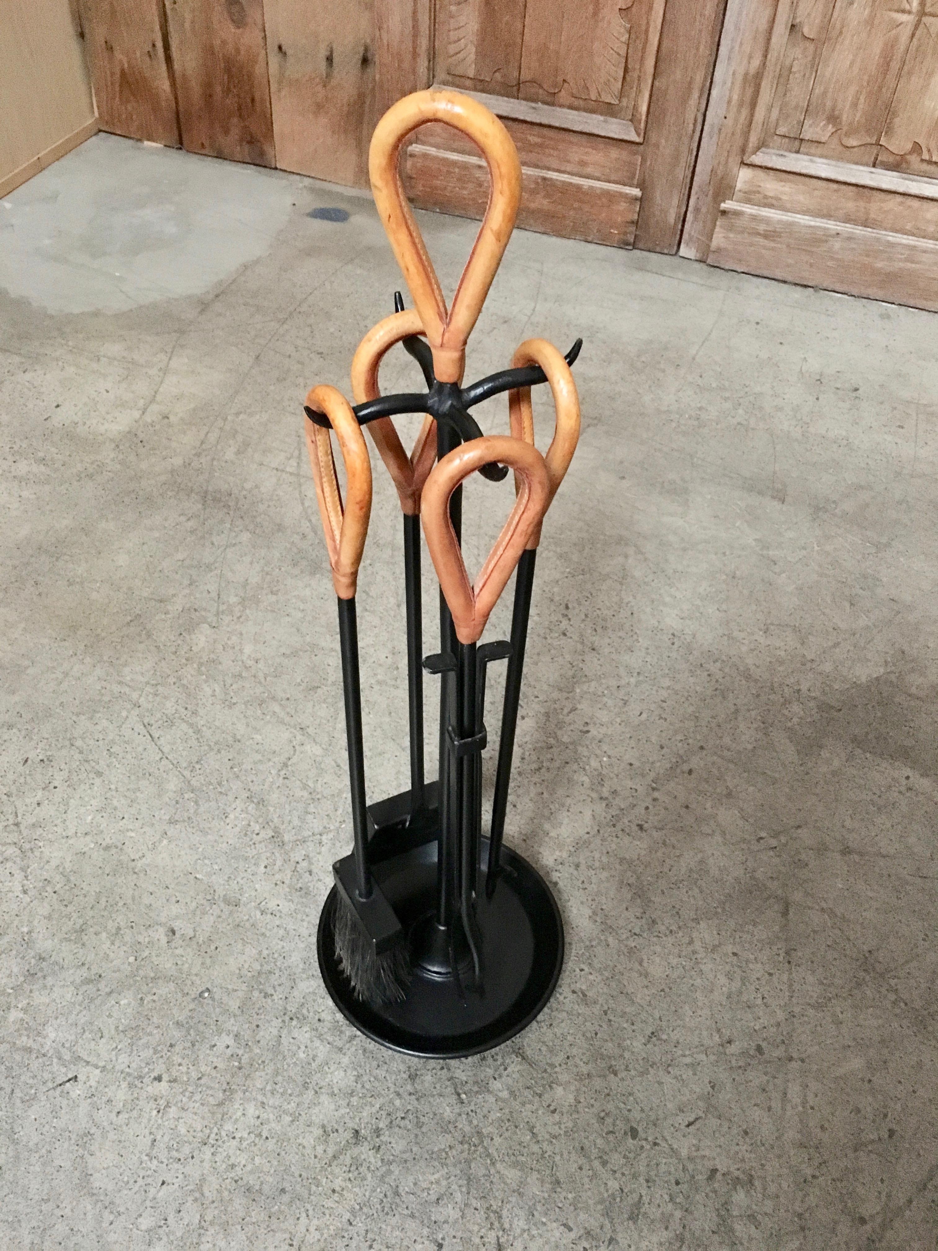 Fireplace set in the style of Jacques Adnet. Iron frame with saddle leather wrapped handles. This fire tool set includes a stand, long tongs, poker, brush and shovel.