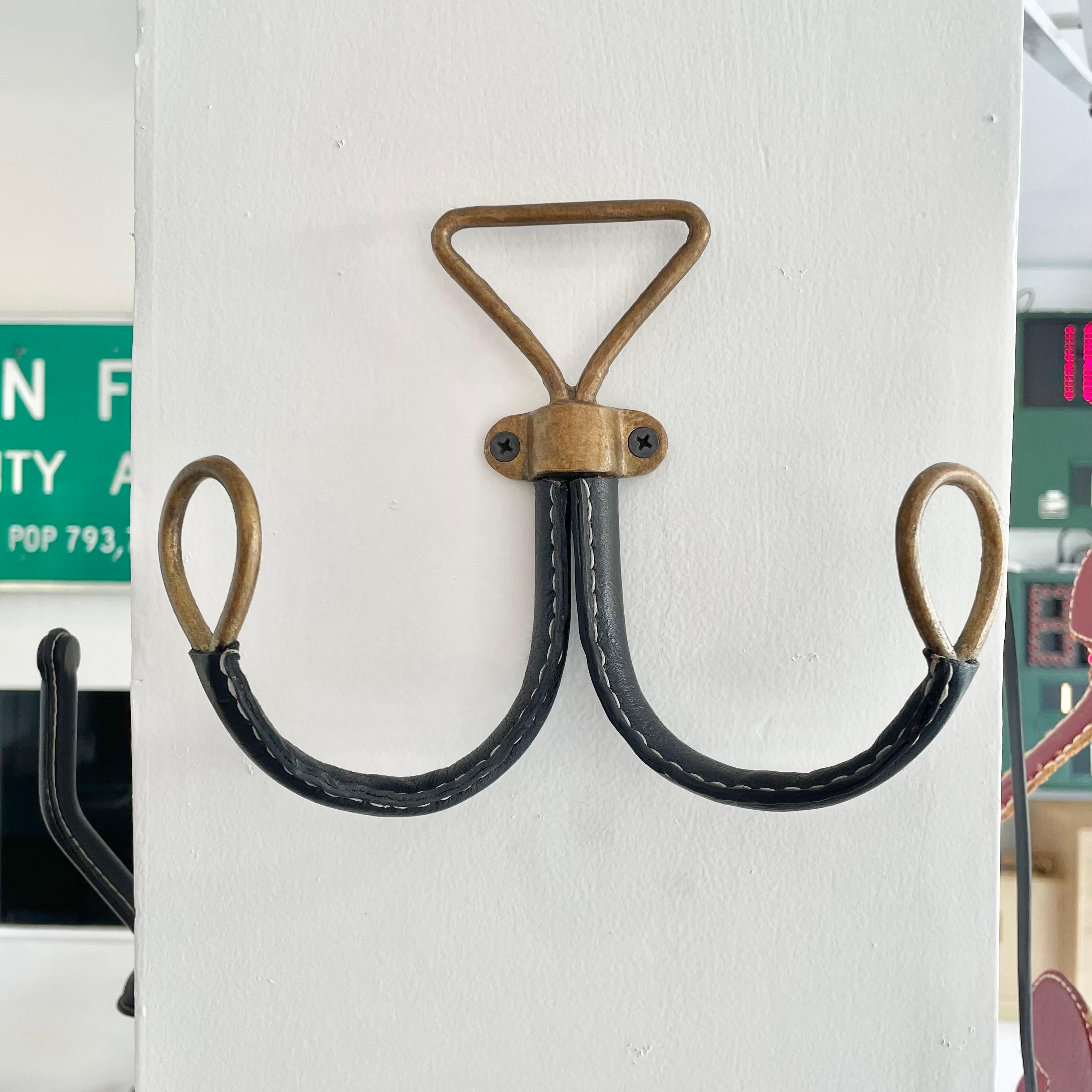 Handsome black leather and brass dual wall hook in the style of French designer Jacques Adnet. Frame made of solid brass and with both arms wrapped in black leather. Each seam is stitched with a contrasting stitch. Held on to the wall by two screws.