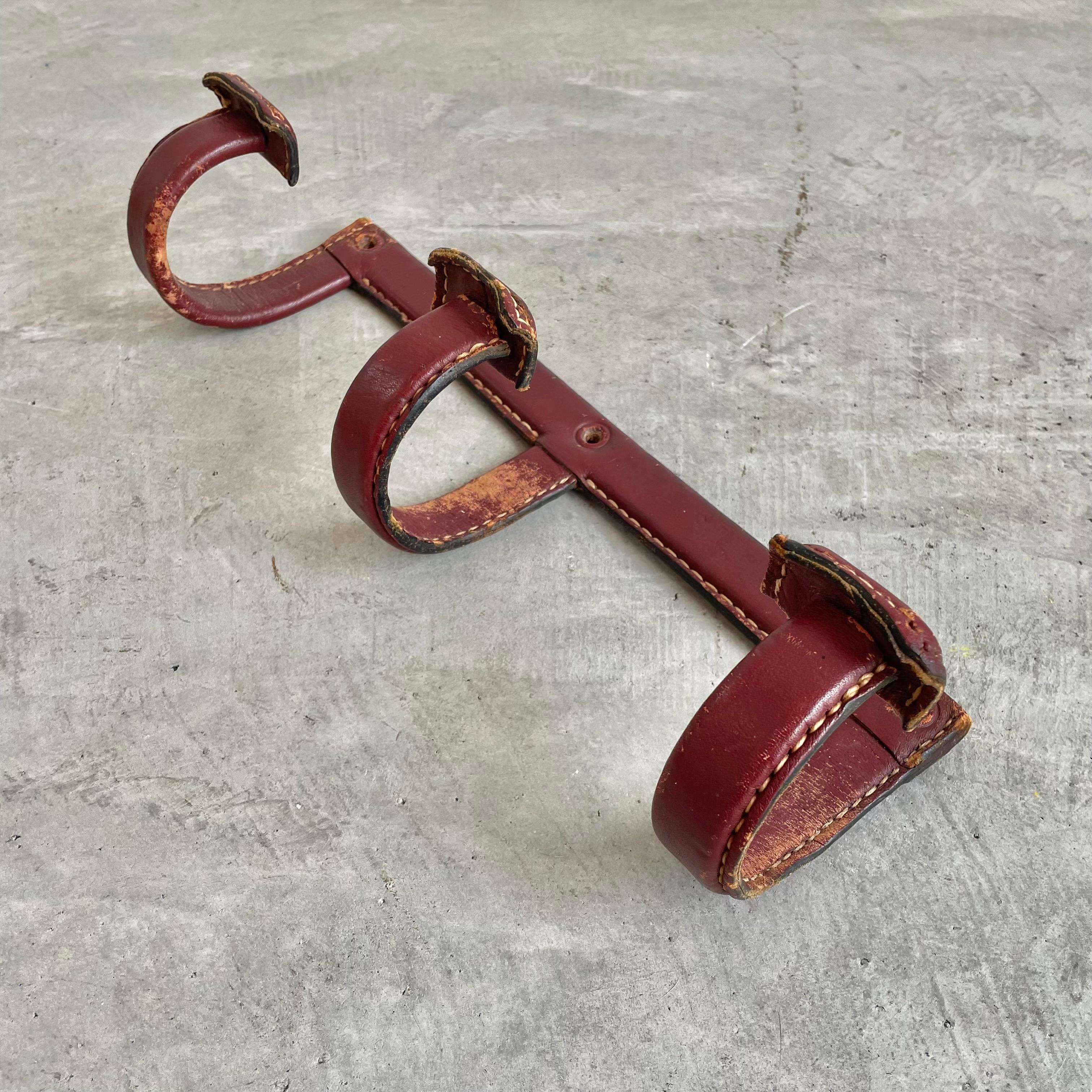 Handsome triple hook coat rack by French designer Jacques Adnet.  Finished in a beautiful oxblood leather with light patina and fading. Frame made of solid iron and completely wrapped in leather. Every seam is stitched with Adnet's signature