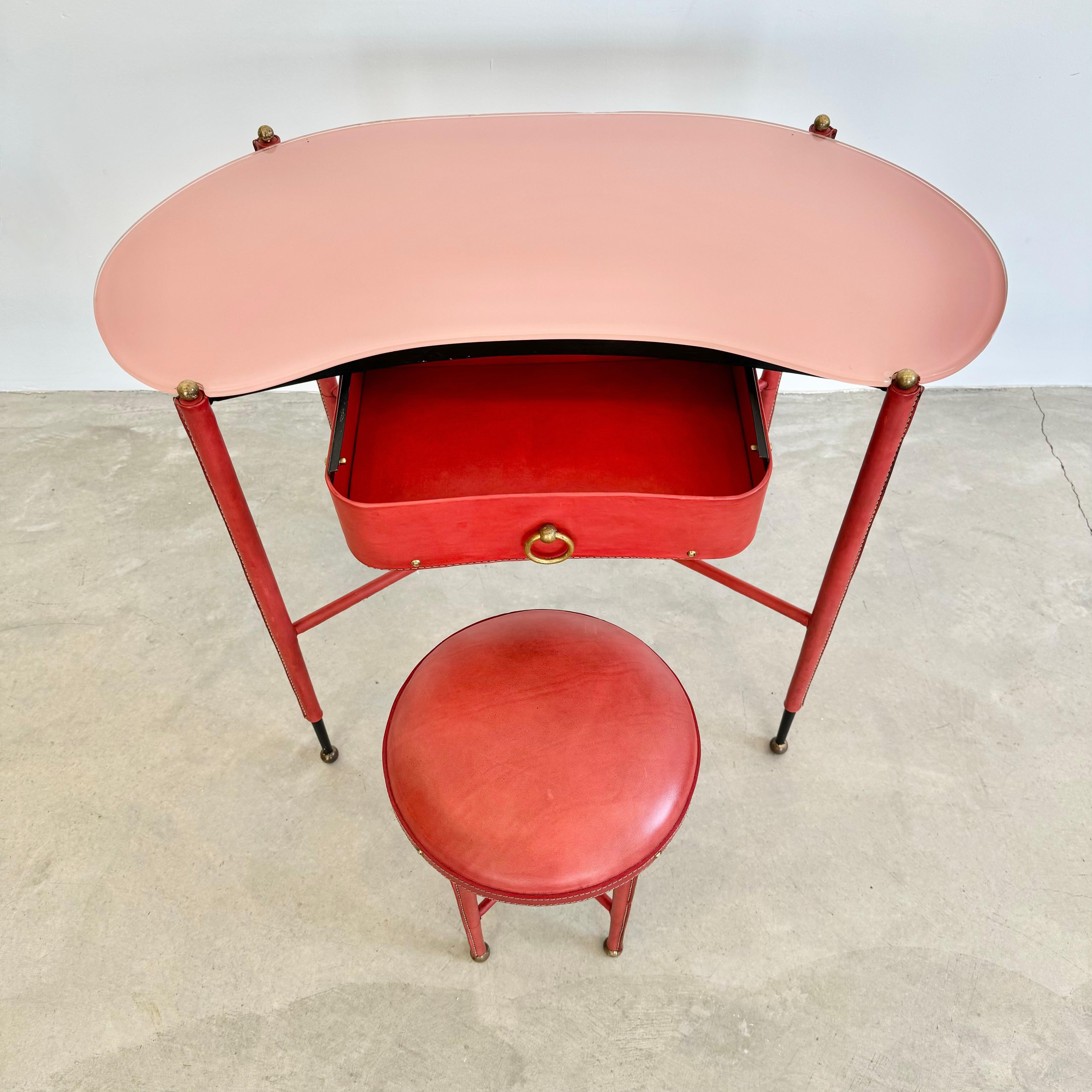 Stunning vanity by French designer Jacques Adnet. Iron frame completely wrapped in red leather. Legs taper down to sharp exposed iron and end with brass ball feet. Tops of each leg is also topped with a brass ball which holds glass table top in