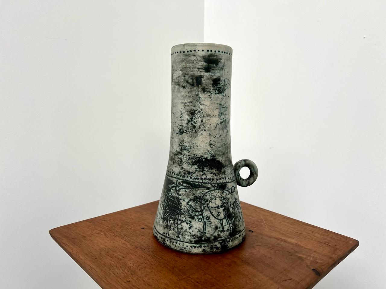 Vaso grande con un piccolo manico staccato, svasato verso il basso. Condizioni perfette. Firma incisa sotto la base. Grigio sbiadito fino al nero, con lievi riflessi verdi in alcuni punti.

La decorazione è un fregio rotante composto da 2 pecore e 2