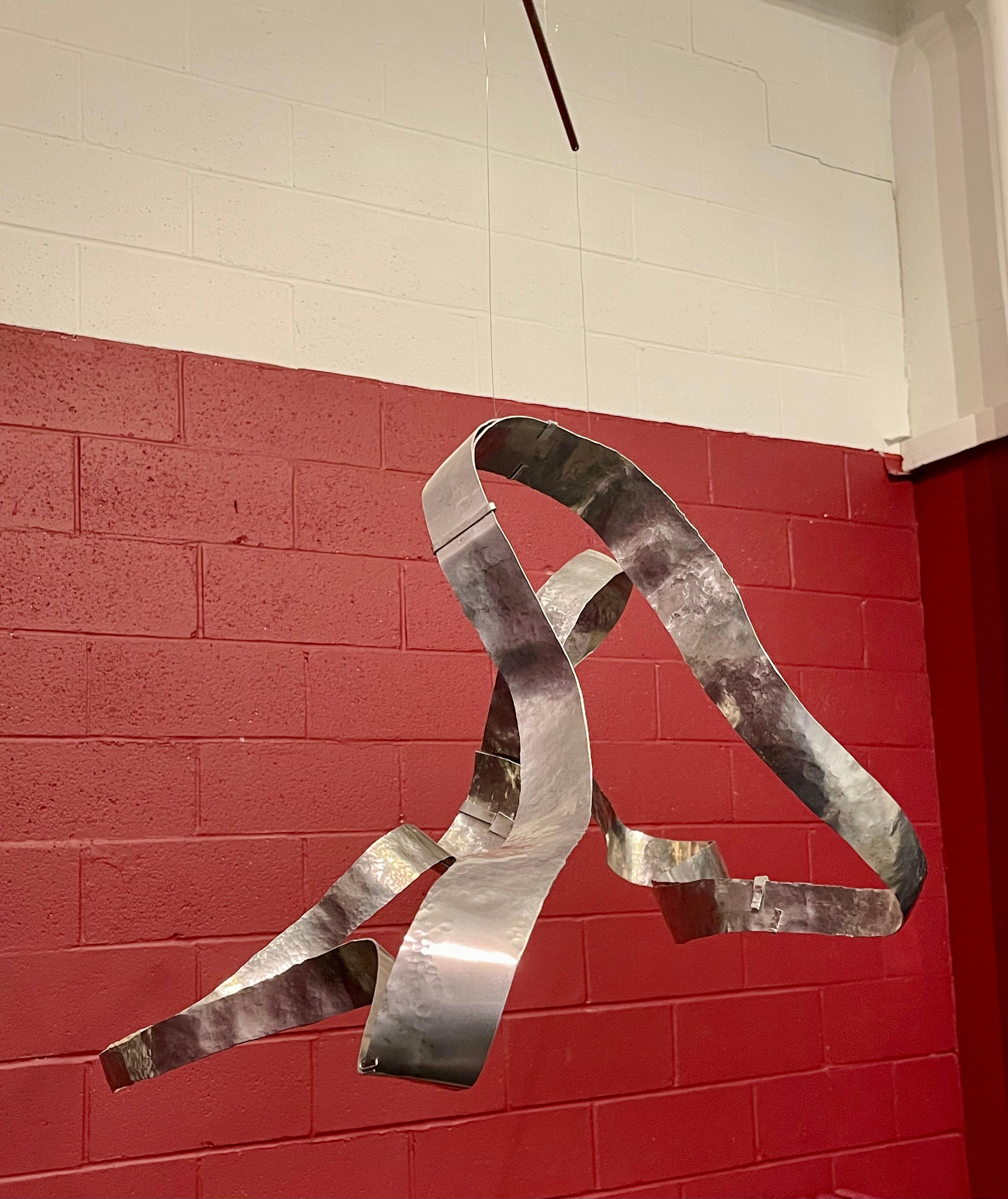 Ola es una escultura suspendida, martillada a mano, que se mueve suavemente con el aire ambiente, entablando con el espacio circundante un diálogo silencioso y continuo. Al girar lentamente, sus contornos cambiantes trazan corrientes invisibles,