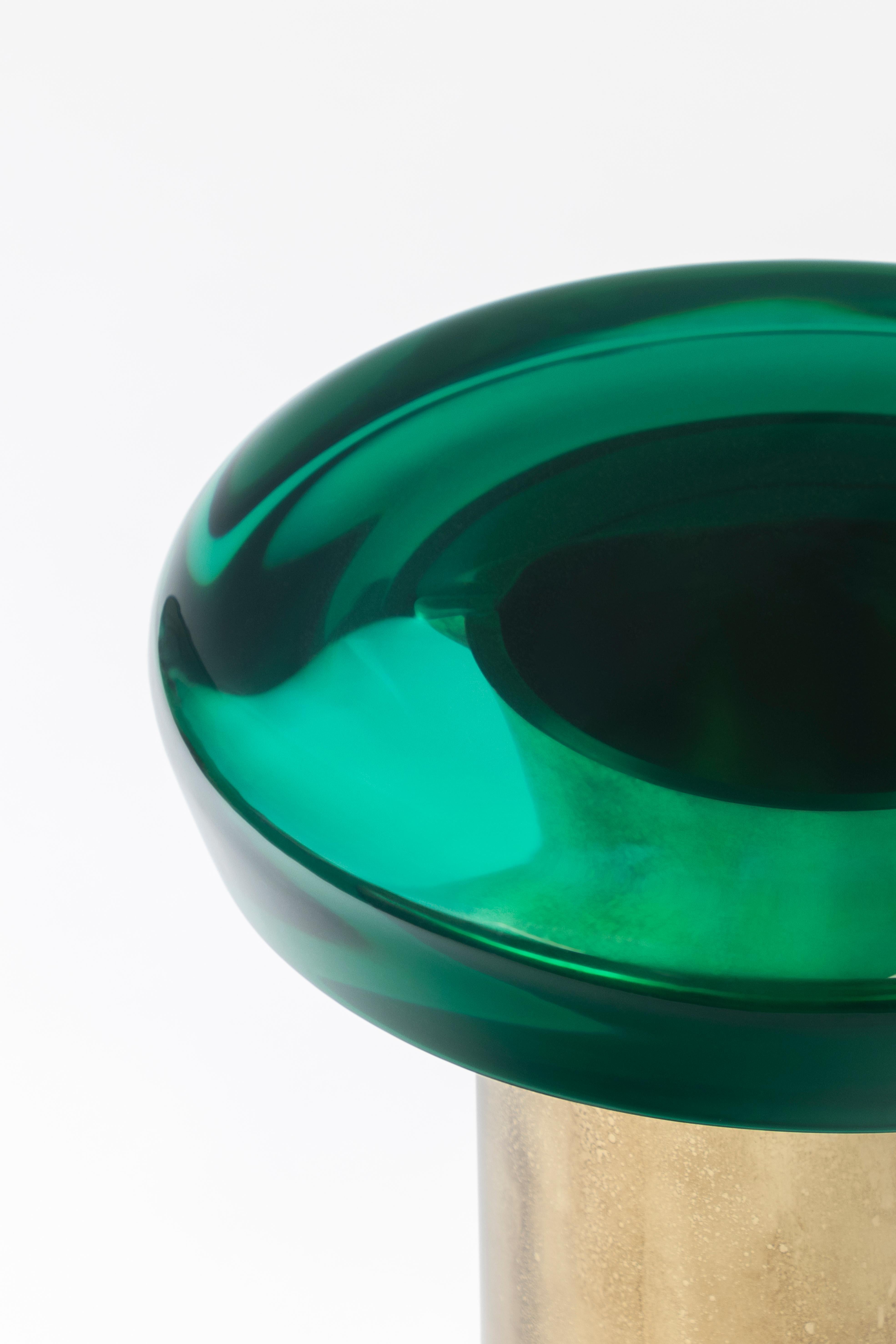 La table basse Jade se caractérise par une transparence éthérée et une surface réfléchissante qui joue naturellement avec la lumière. Le plateau de table aux couleurs vives repose sur une base cylindrique en laiton, dont les veines distinctives sont