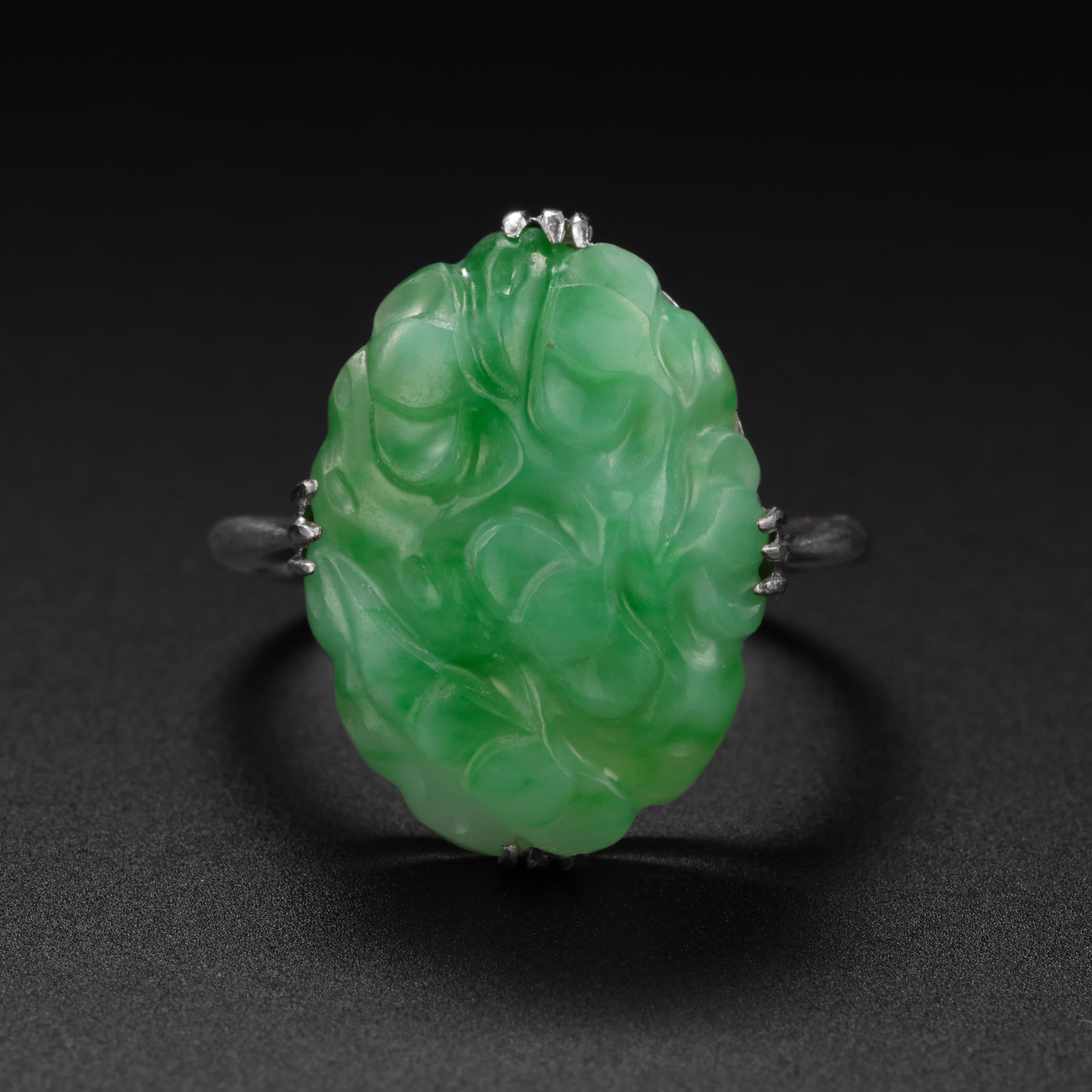 This 1920s-1930s Art Deco ring was hand-crafted in 18K white gold and features a luscious, translucent bright apple-green oval jadeite jade carving.  The luminous stone is deeply carved and measures 17.0vmm 12.73mm x 3.98mm. The foliate design of