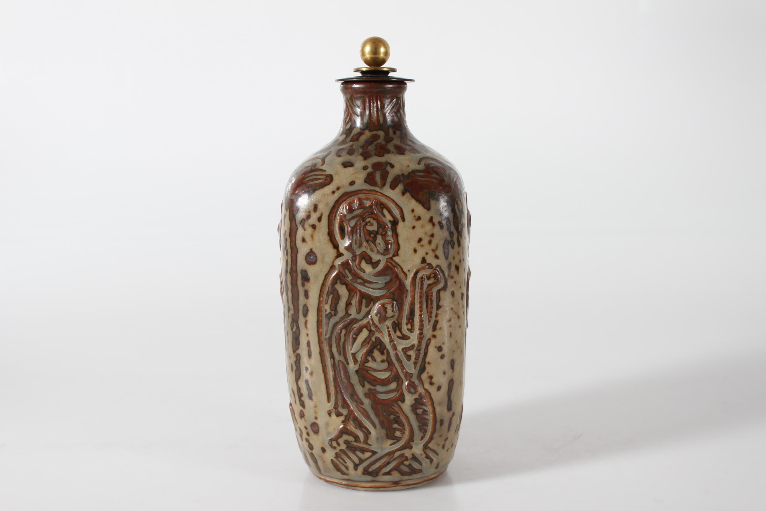 L'artista danese Jais Nielsen ha disegnato questo sottile vaso o bottiglia quadrata in gres con coperchio in bronzo per Royal Copenhagen.

I quattro lati mostrano una decorazione di motivi biblici ricoperti di smalto Sung An marrone e beige. Il tema