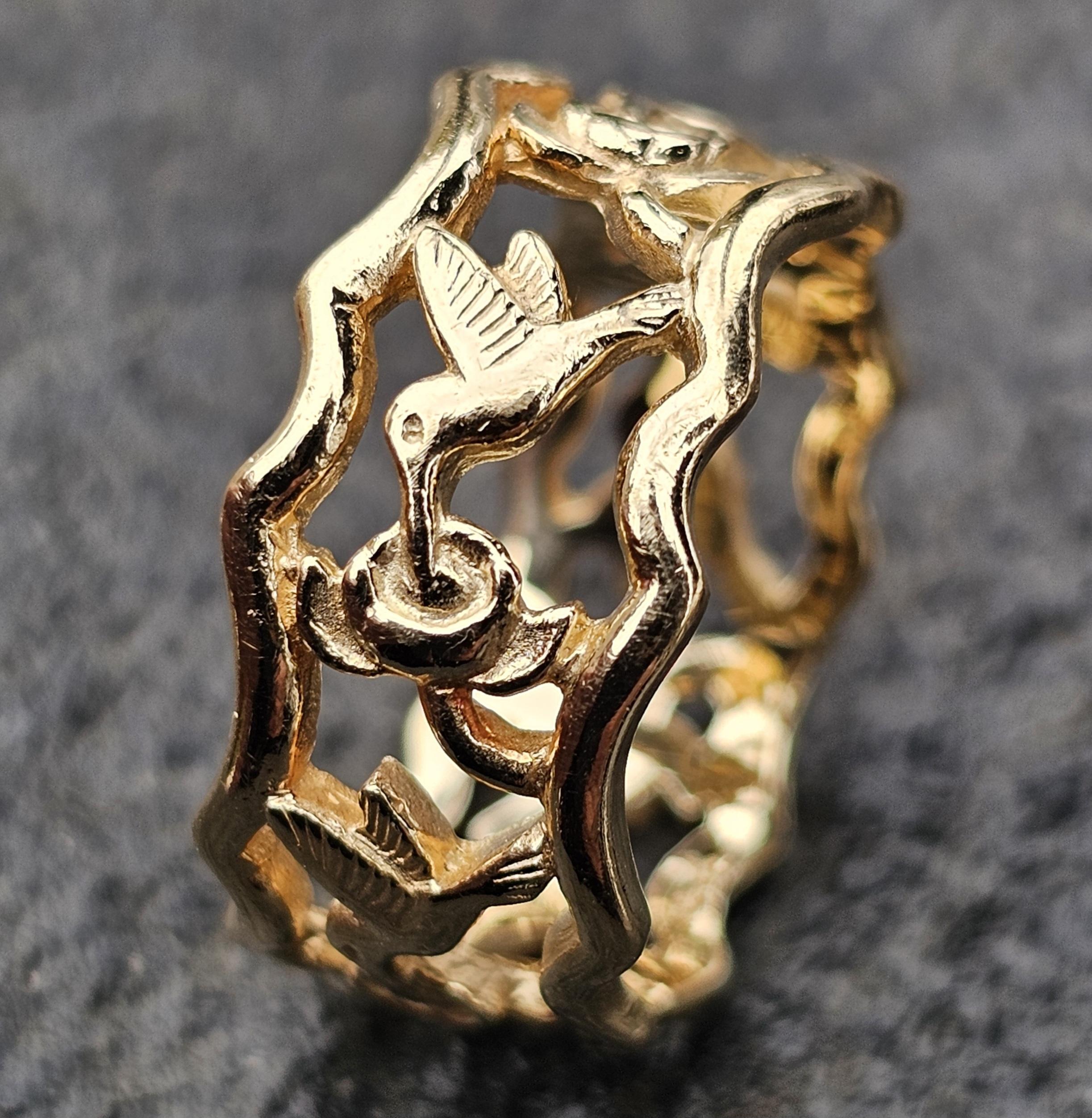 Questo raro anello a fascia di James Avery ritirato è un pezzo accattivante di design moderno. Realizzato in oro giallo 14k, presenta un intricato motivo traforato con colibrì e fiori. L'anello è una misura 7,5, pesa 5,9 grammi e presenta una