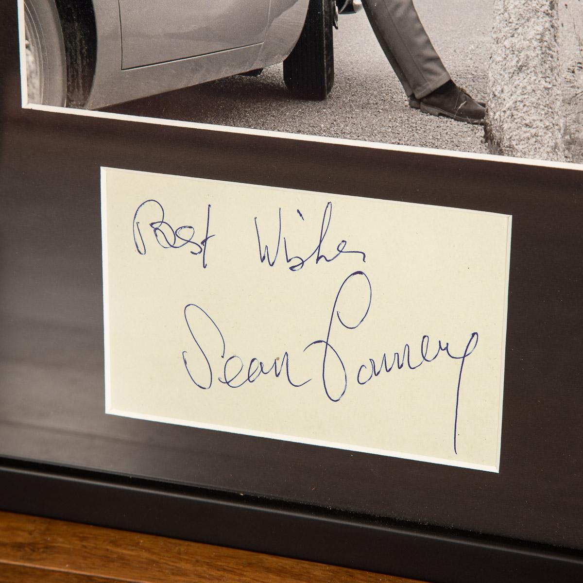 Schwarzweißfotografie von Sean Connery als '007' mit dem 'Goldfinger' Aston Martin DB5 am Furkapass, 24,5 x 19,5 cm, Display montiert mit Unterschrift von Connery in blauer Kugelschreiberschrift, mit Widmung 