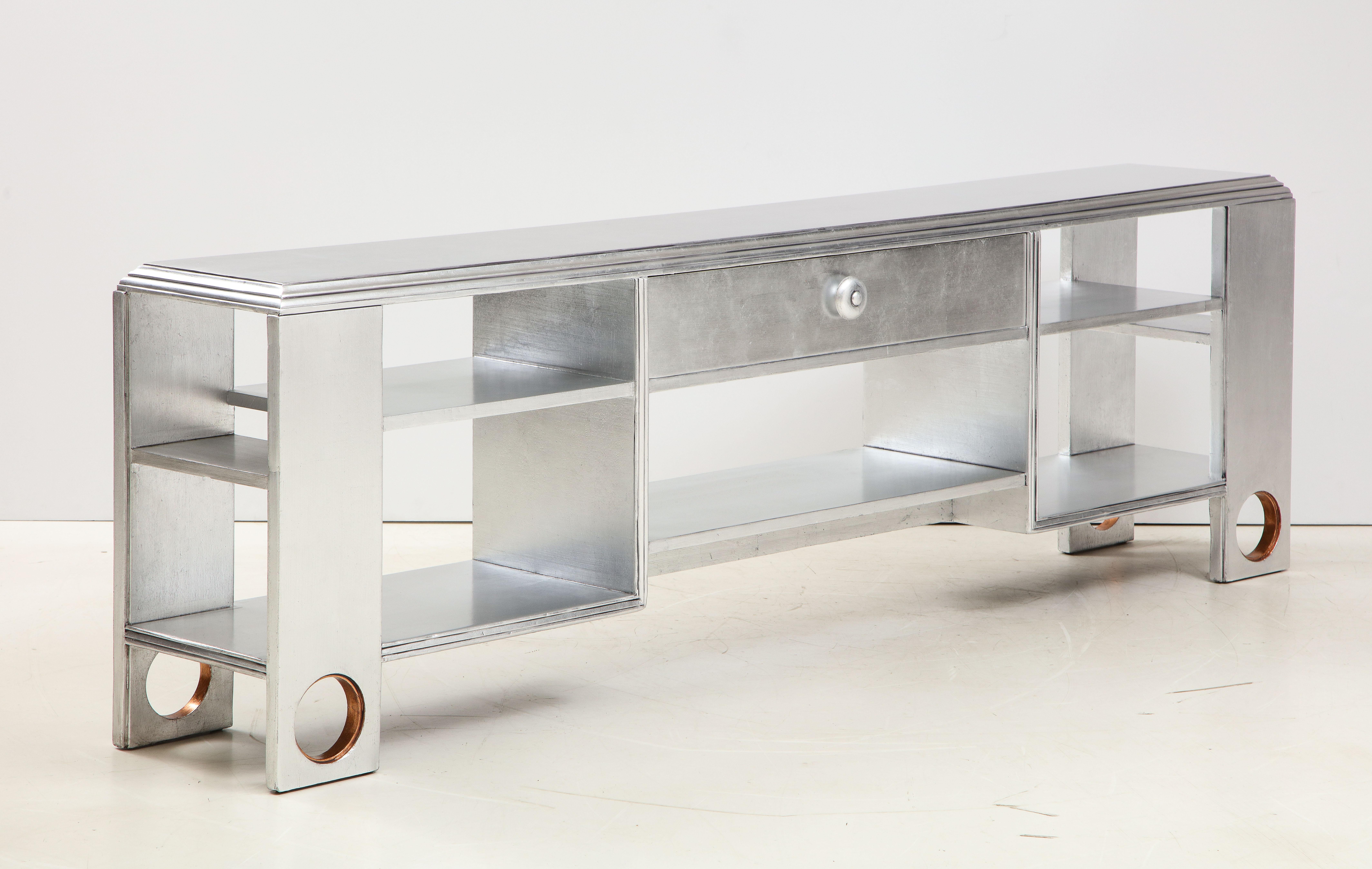 Majestic mid century console by James Mont featuring a hand applied glazed silver leaf custom finish. Console has 3 longer shelves and center drawer and 2 hidden side shelves. Each of the four supports feature a circular cutout which jave a