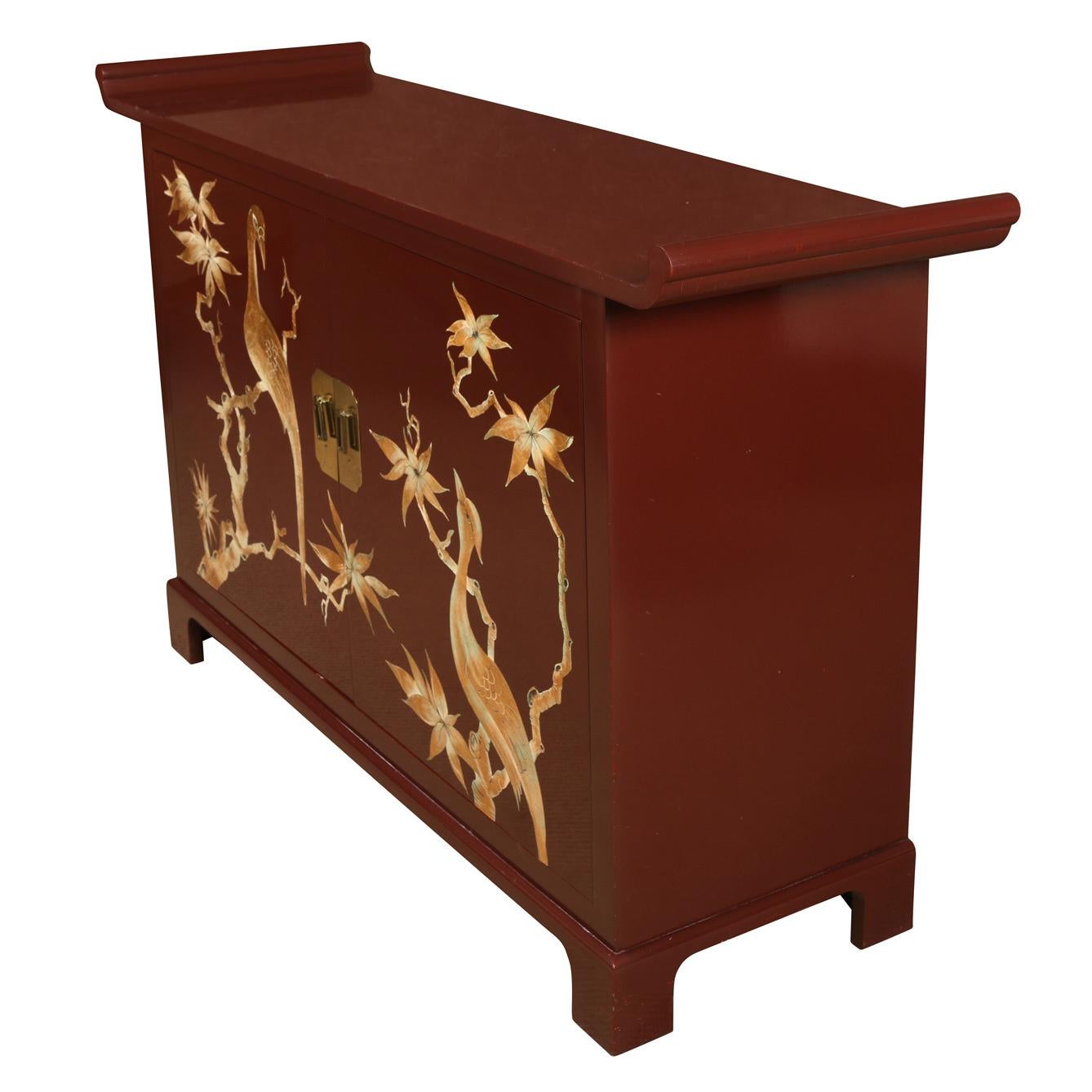 A vintage James Mont style, Asian inspired,  sideboard in a wine hue with brass details, depicting birds, tree branches and leaves.  Two doors open to reveal two large shelves for storage