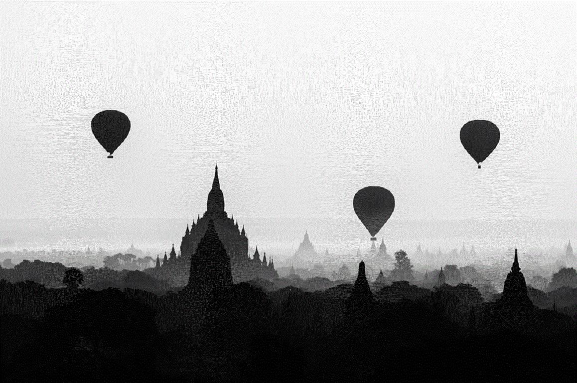 Kurz nach Sonnenaufgang löst sich der Nebel auf und gibt den Blick auf ein Panorama von Pagoden frei, das sich über das Tal von Bagan in Birma erstreckt.  Eine Flottille von Heißluftballons, die langsam in der kaum wahrnehmbaren Brise dahintreiben,