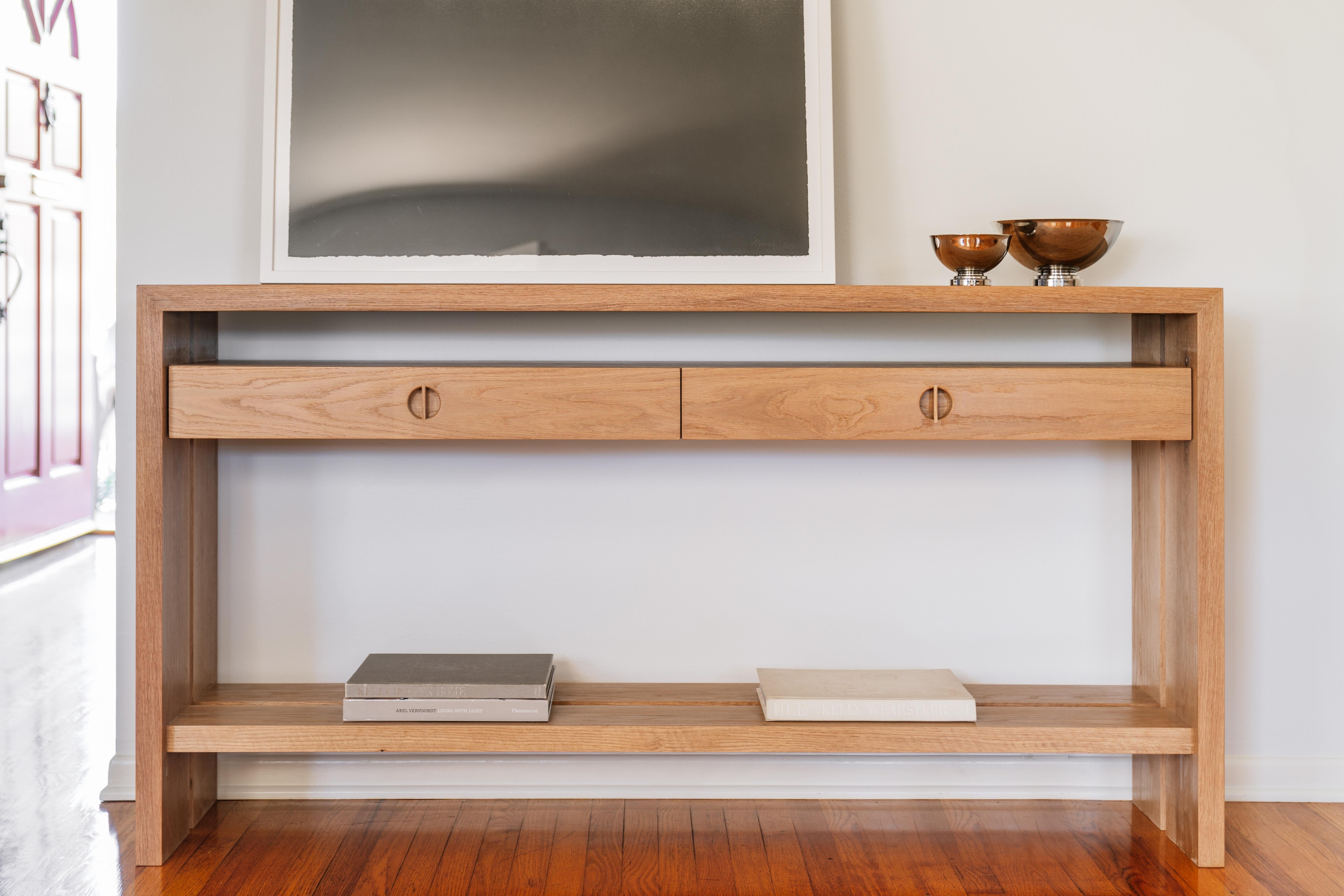 This custom wood console table is hand-made in the United States with all hardwood construction. It features a modern design with a solid white oak body and a minimalist walnut inlay detail. Convenient and accessible storage drawers feature