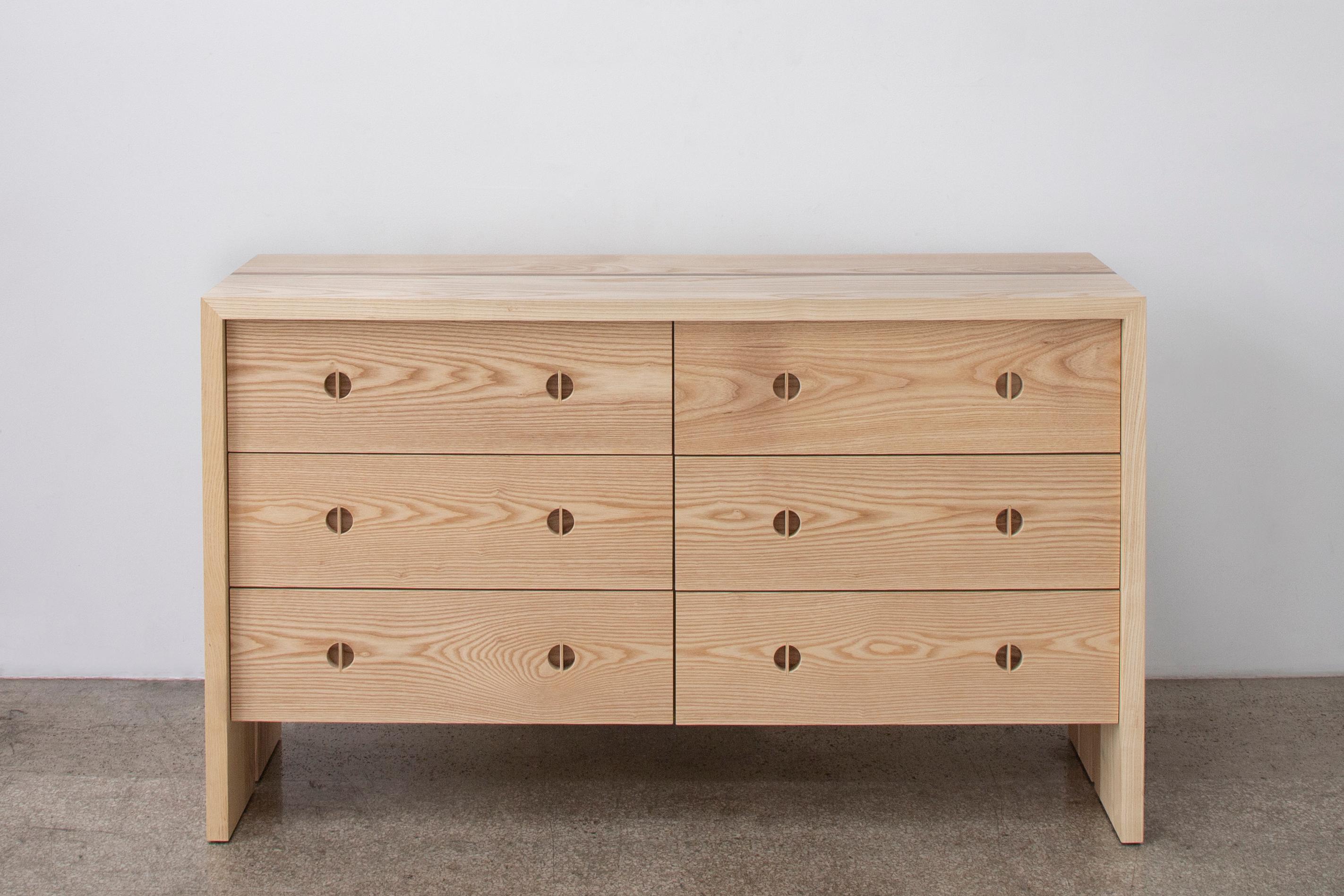 Questo comò moderno in legno è realizzato a mano negli Stati Uniti con una struttura interamente in legno duro. Caratterizzato da un corpo in frassino massiccio, da un intarsio minimalista in noce e dall'esclusivo dettaglio delle gambe a spacco,
