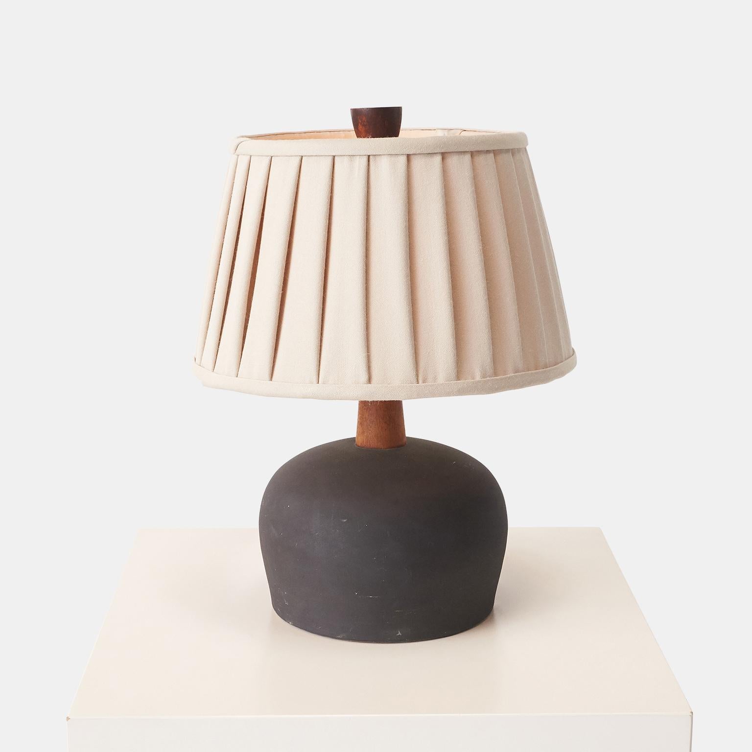 A matte black table lamp by Jane and Gordon Martz for Marshall Studios. Stoneware base with teak neck and finial.

Complete with a beautiful hand-crafted pleated shade made from a luxurious Sandra Jordan Prima Alpaca quinoa colored fabric from the