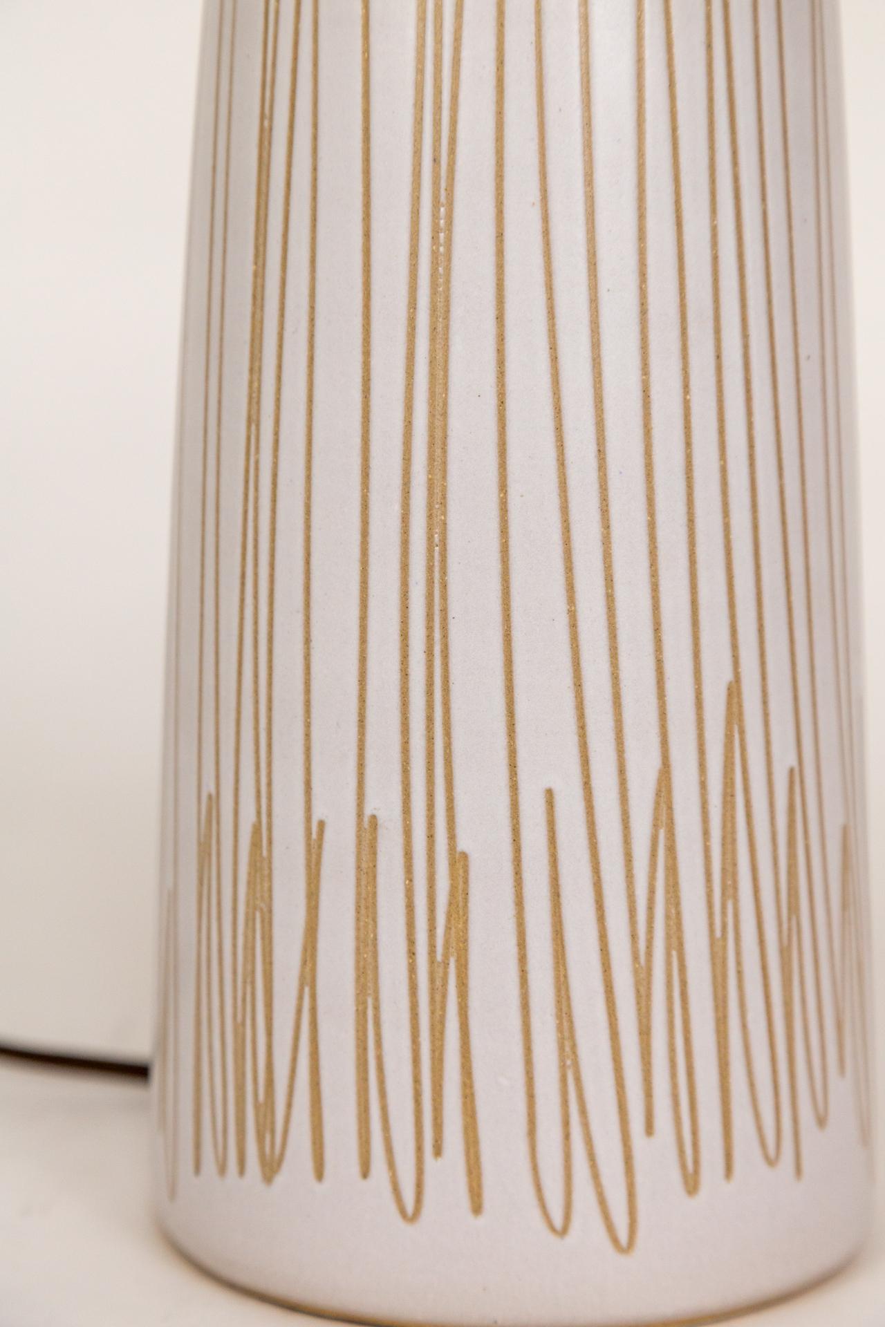 A streamlined, ceramic lamp in white satin glaze with scribbles revealing the raw clay; made by Marshall Studios and designed by Jane and Gordon Martz, circa the 1960s.  It is cylindrical shape with a cherry stained wood neck. Signed on the back. 