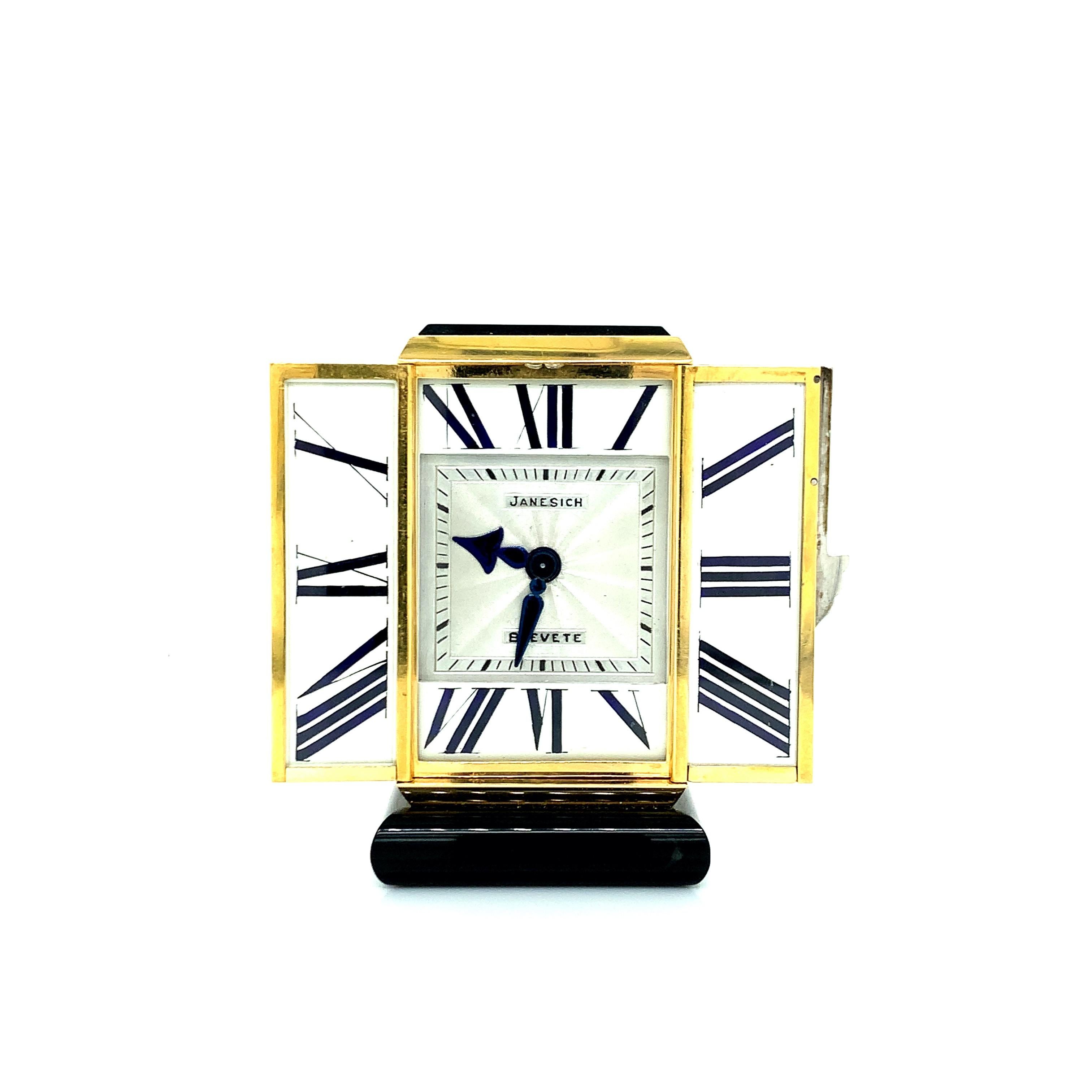 This travel clock is triptych shaped. When its doors are opened, the dial appears, which is adorned with hours in black enameled Roman numerals on a white background under glass. Square dial in silver guilloché with railroad and blued steel hands.