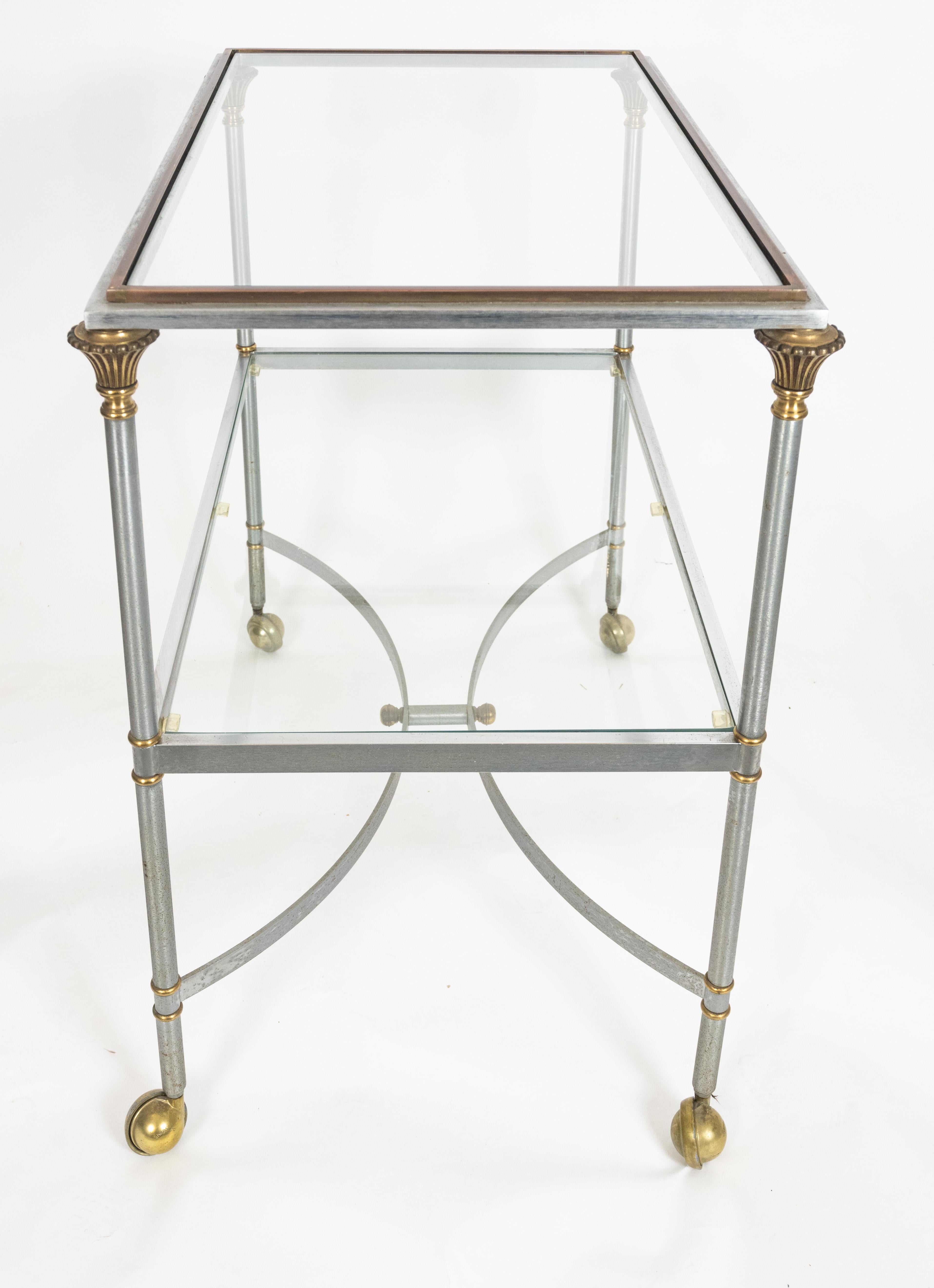 A two-tiered rectangular cart in the manner of Jansen Maison with glass surfaces inset with a steel and bronze frame. The four legs joined by c-shaped stretchers and supported by round brass castors. Circa 1960.