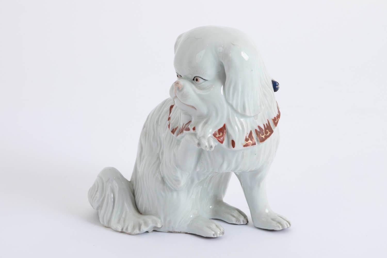 Small sculpture of white Kutani porcelain with enameled decoration representing a sat dog with long hair, the head turned to the right and the right paw raised. It wears a blue necklace with yellow, green and red motifs, on which square red pendants