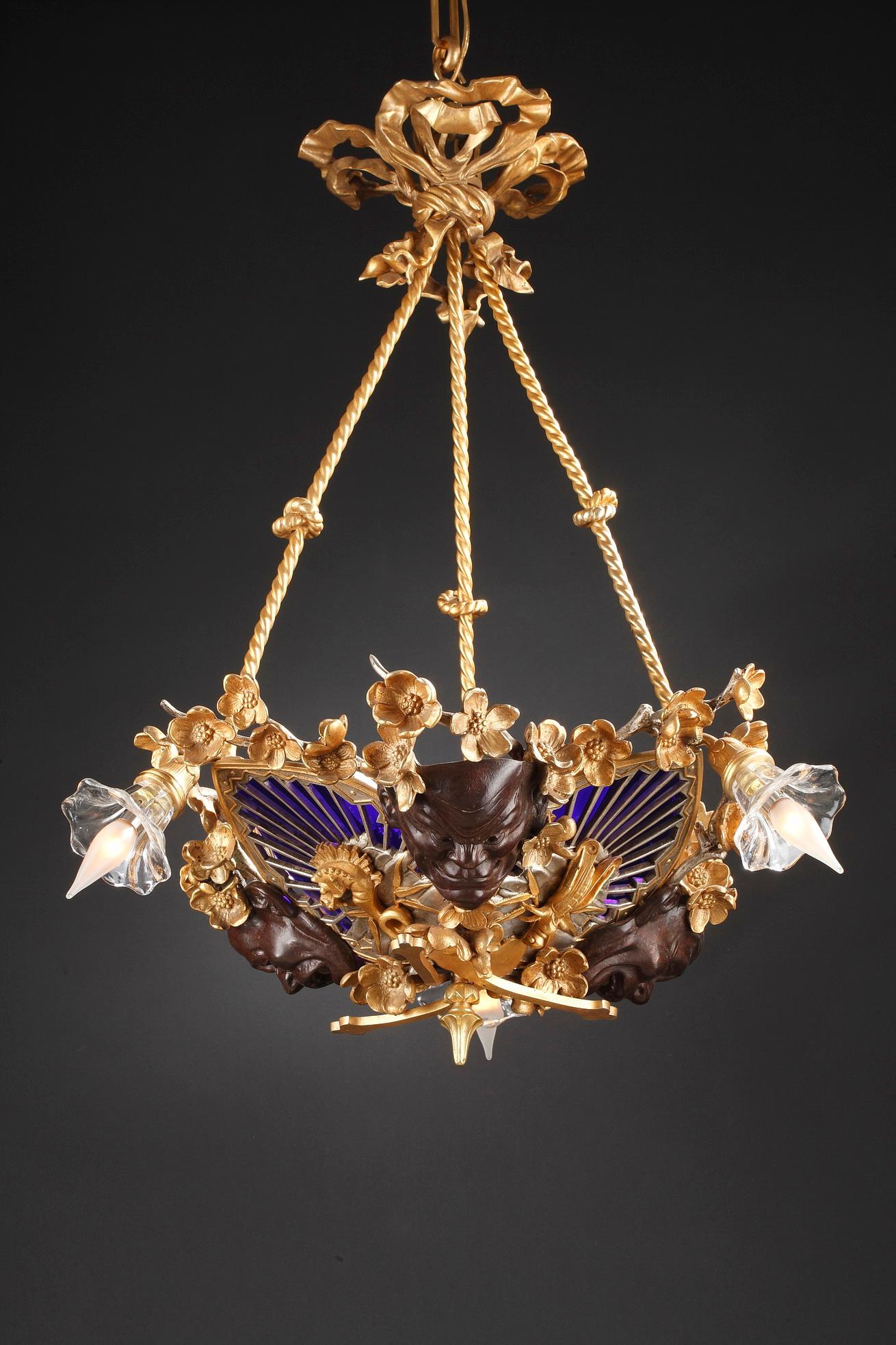 Beautiful Japanese style chandelier attributed to E. Soleau in gilded, silvered and patinated bronze with three light-arms ending with corollas. It is composed of three patinated bronze masks inspired by the Nô theater, and three stylized fans,