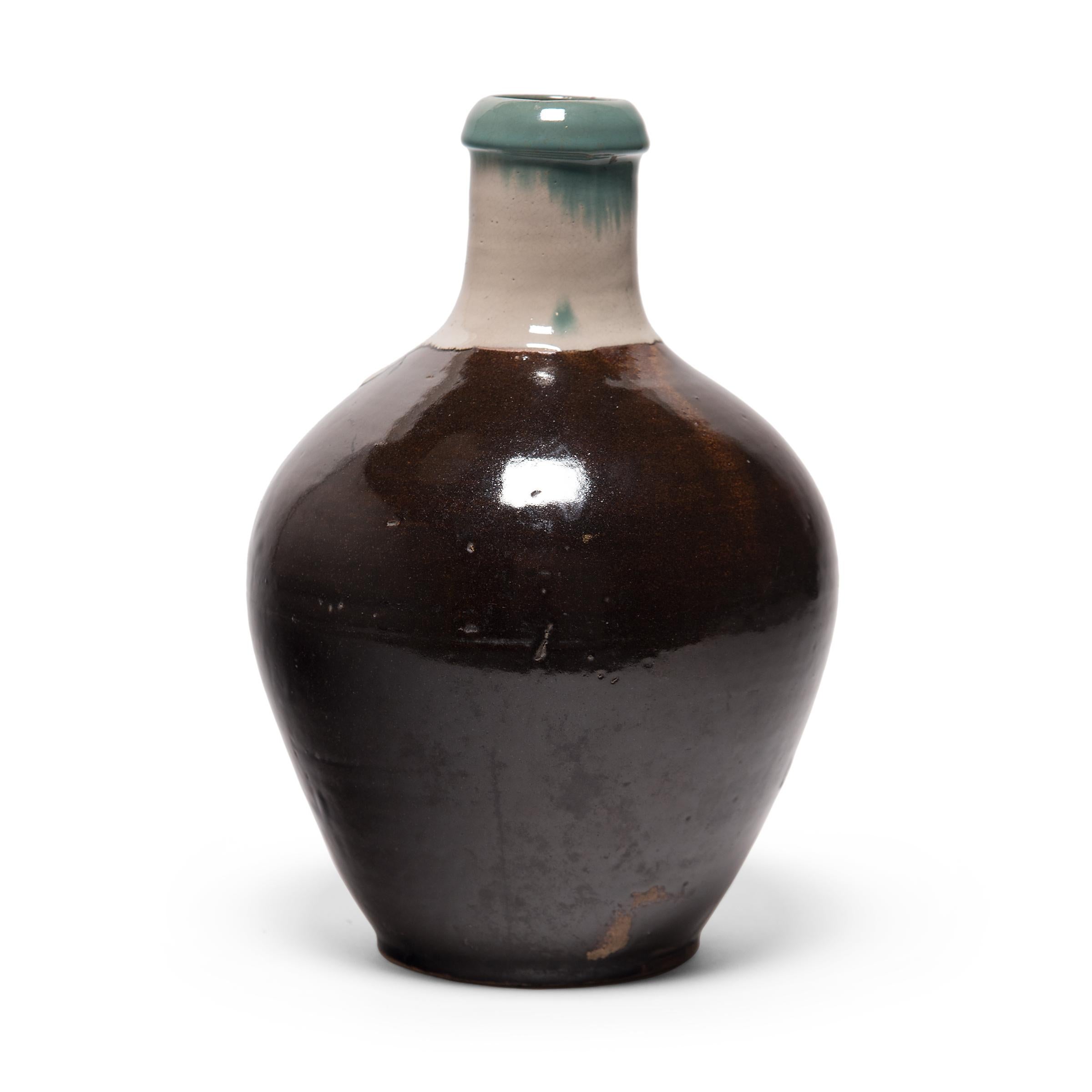 This unusual bottleneck jar is a late-Meiji era Japanese sake bottle, also known as tokkuri. Used to heat and serve sake, the ceramic bottle has a graceful, tapered form with wide shoulders and an elongated neck for conserving heat. A dark brown