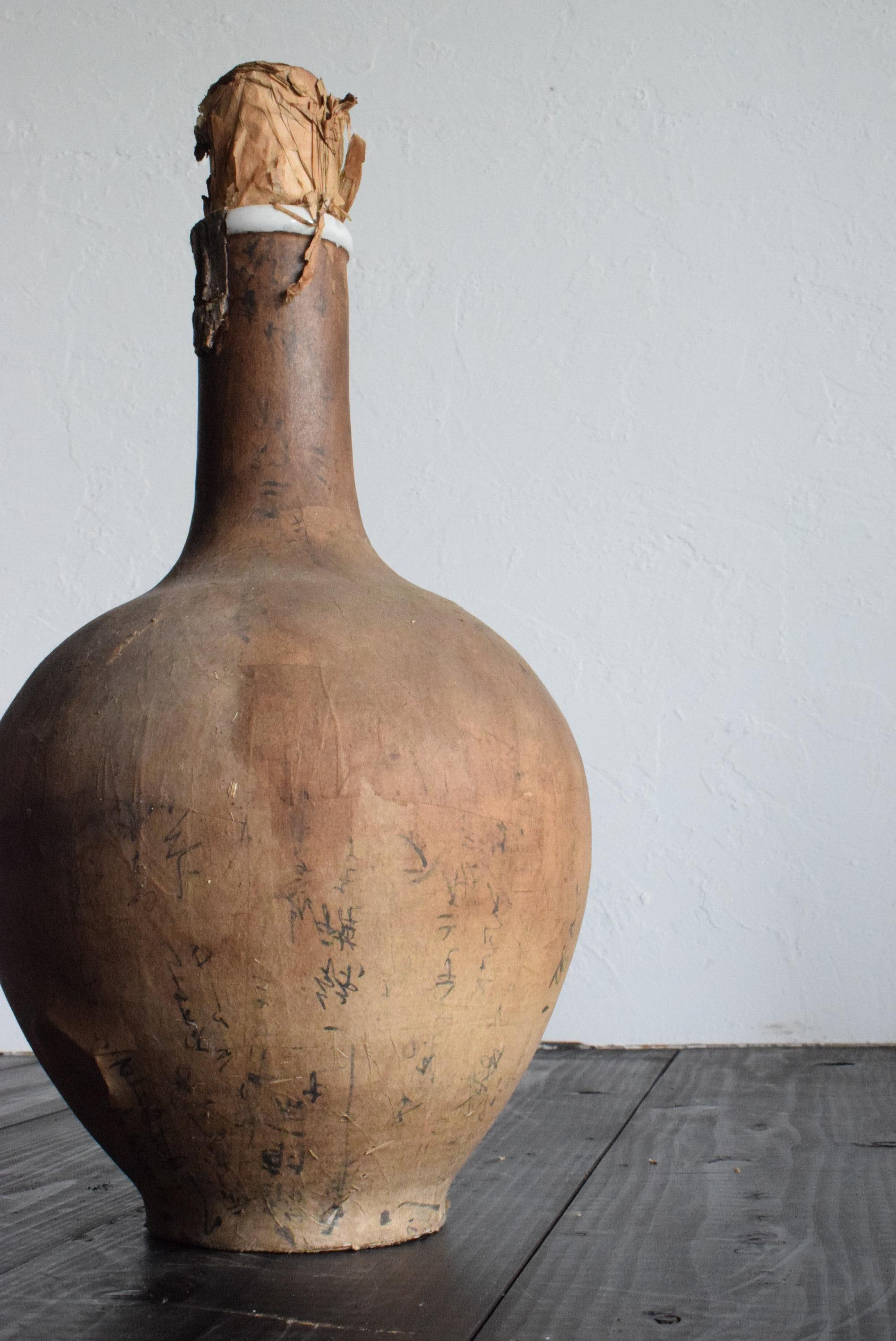 This is an old Japanese sake bottle. It is from the Meiji period (1860s-1900s). It is made of ceramic and covered with paper. It is slightly cracked and reinforced with paper. The paper is peeling off in places, giving it the beauty of an abstract