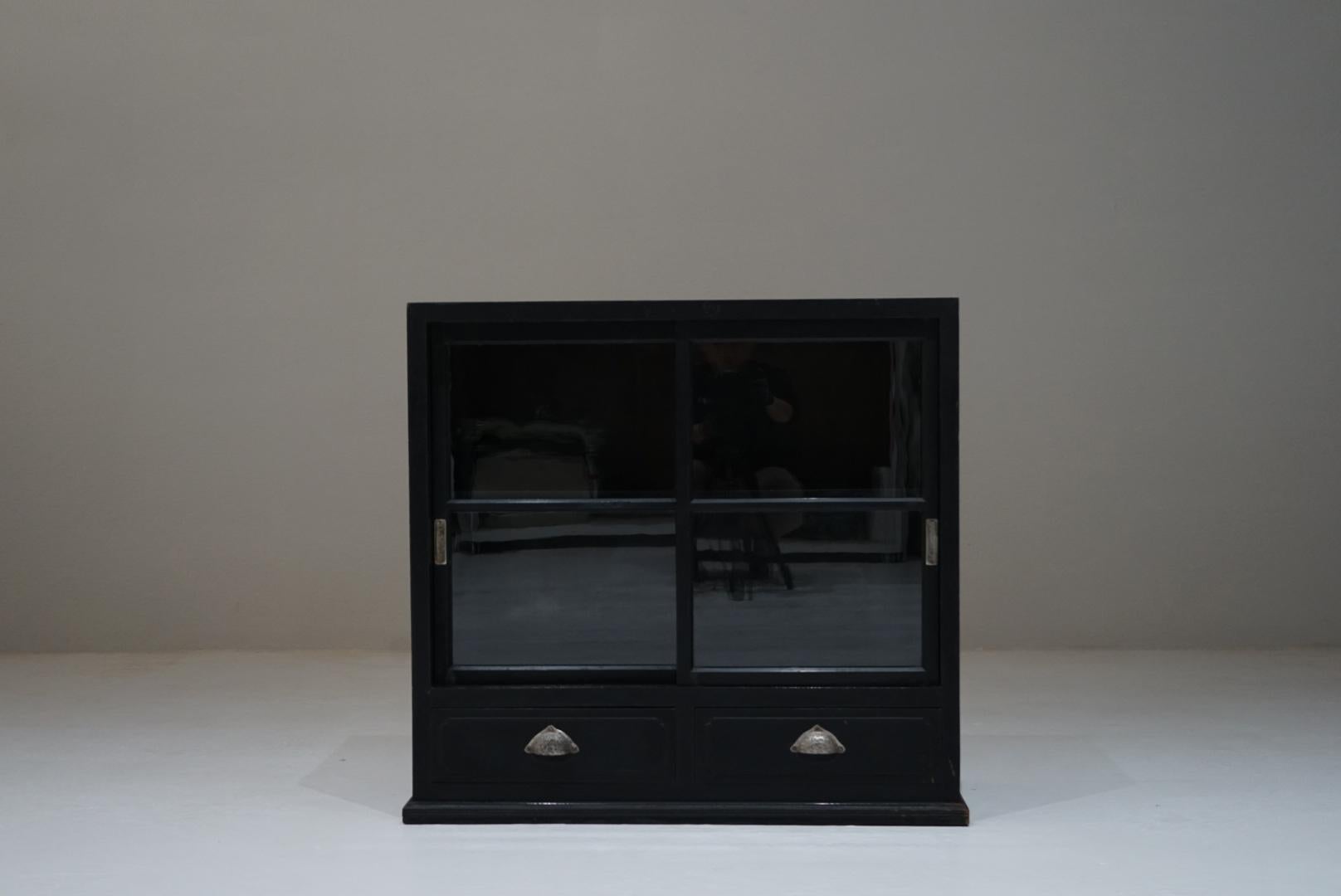 A Japanese antique wooden glass cabinet dating from the 1930s–1950s.
Compact in scale, the piece features small glazed doors that give it a light, approachable presence despite its sturdy construction.
The original black finish from the period