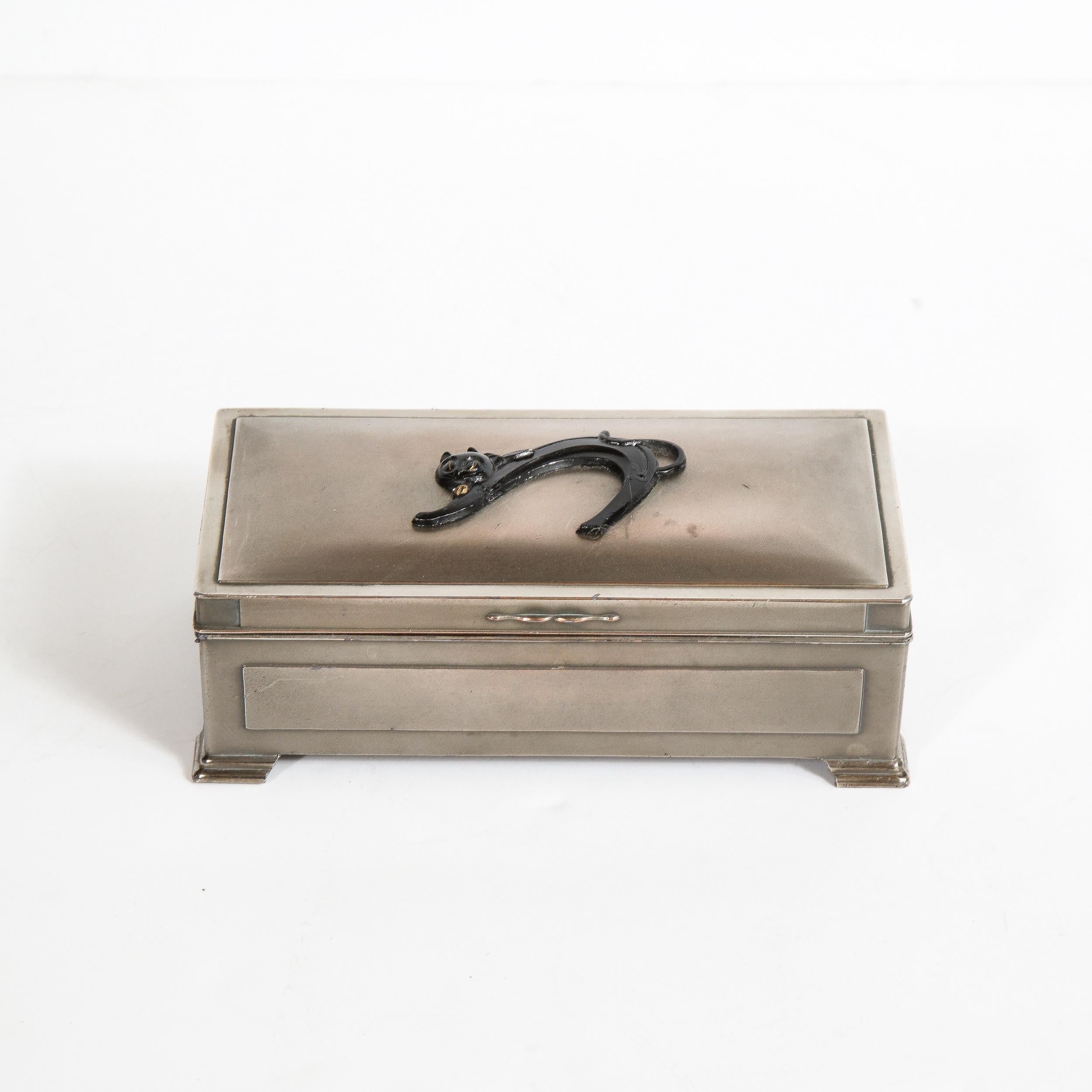 This refined Art Deco pewter box was realized in Japan, circa 1935. It features a volumetric rectangular body with skyscraper style feet; an undulating streamlined handle; and a stylized cat motif in enamel relief adorning the top. Additionally,