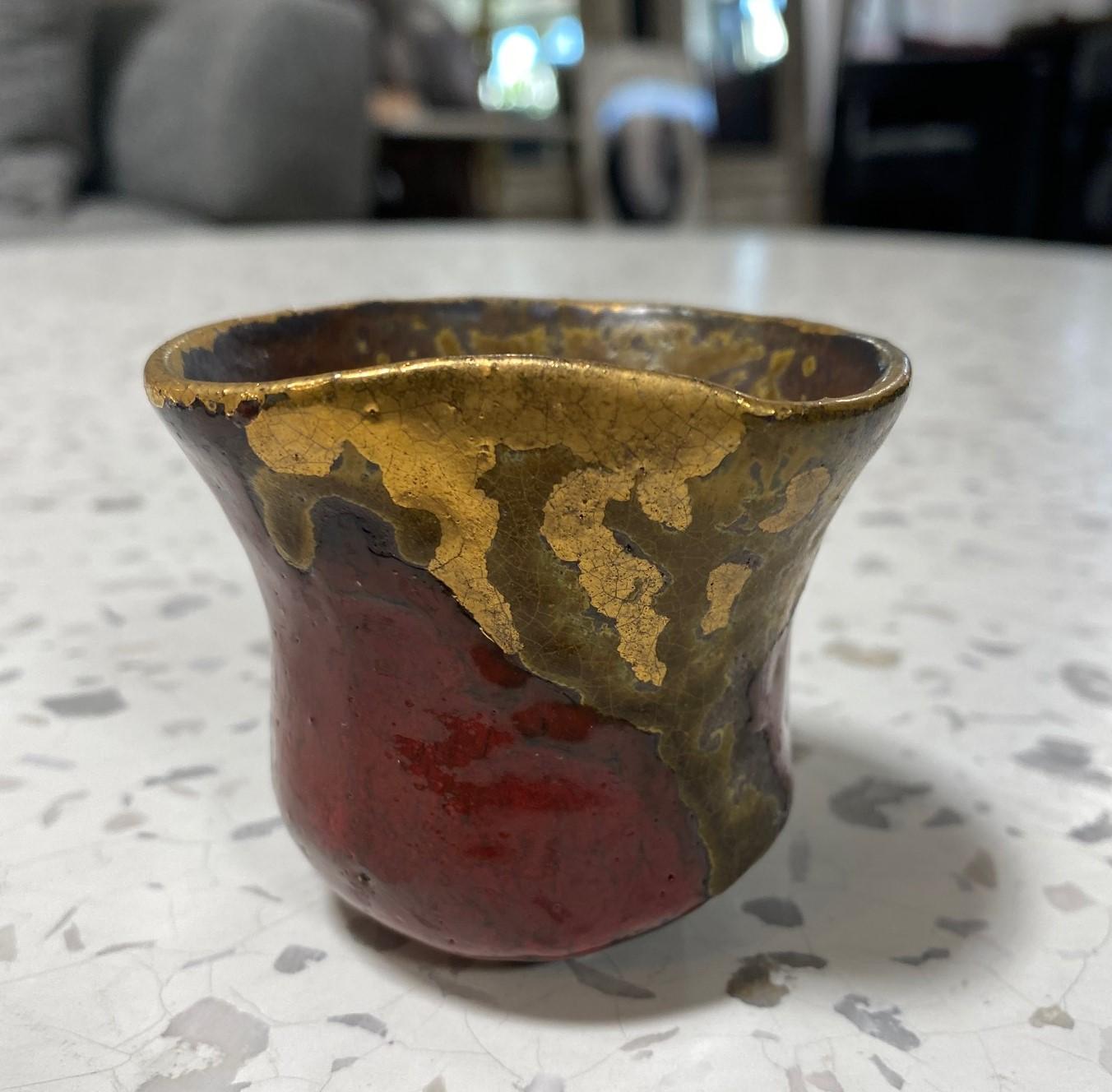 A gorgeous Japanese studio pottery Yunomi teacup or sake cup that features a wonderful sumptuous crimson red glaze with dripping gold gilt glaze and various shifts in color and texture. 

This particular Yunomi encompasses the Japanese Wabi-Sabi
