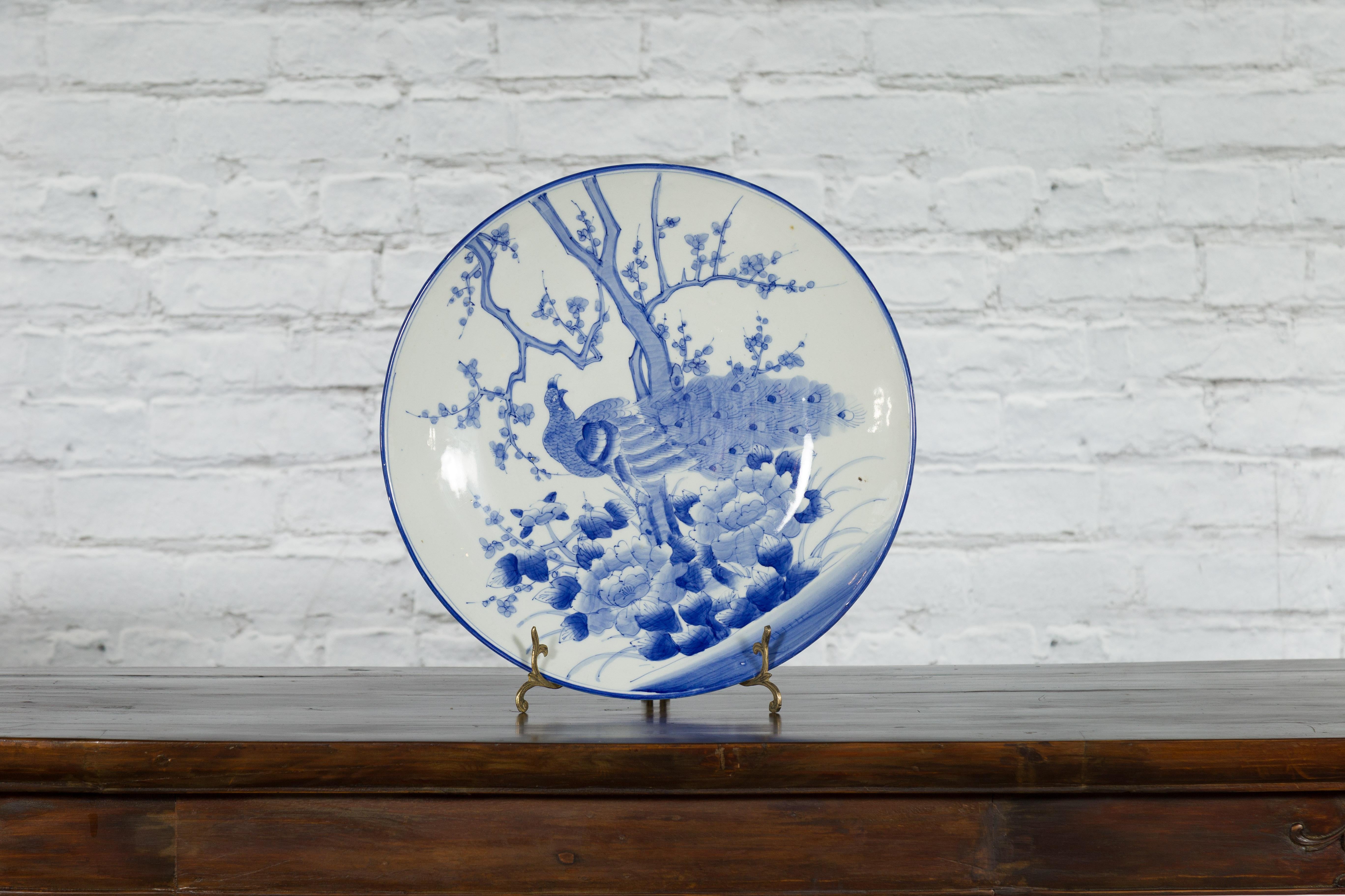 A Japanese porcelain charger plate from the 19th century, with hand-painted blue and white blooming tree, flowers, foliage and peacock décor. Created in Japan during the 19th century, this porcelain plate features a delicate blue and white décor