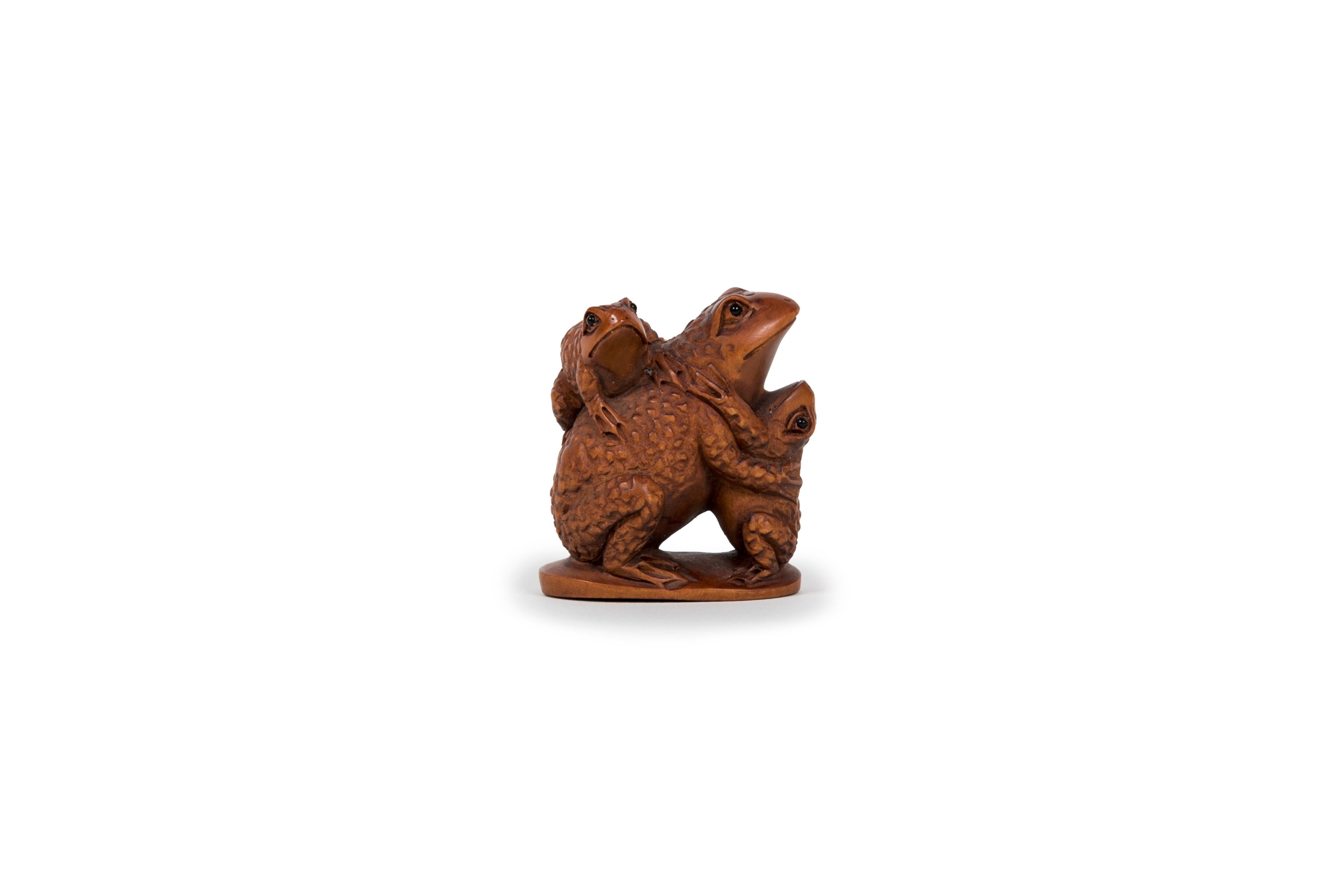 Netsuke in legno di bosso che rappresenta un gruppo di tre rospi, adagiato su una base piatta, occhi intarsiati in corno di bufalo d'acqua. Firmato sotto la piattaforma. La rana (kaeru) è associata alla fortuna e alla ricchezza. Nel Giappone