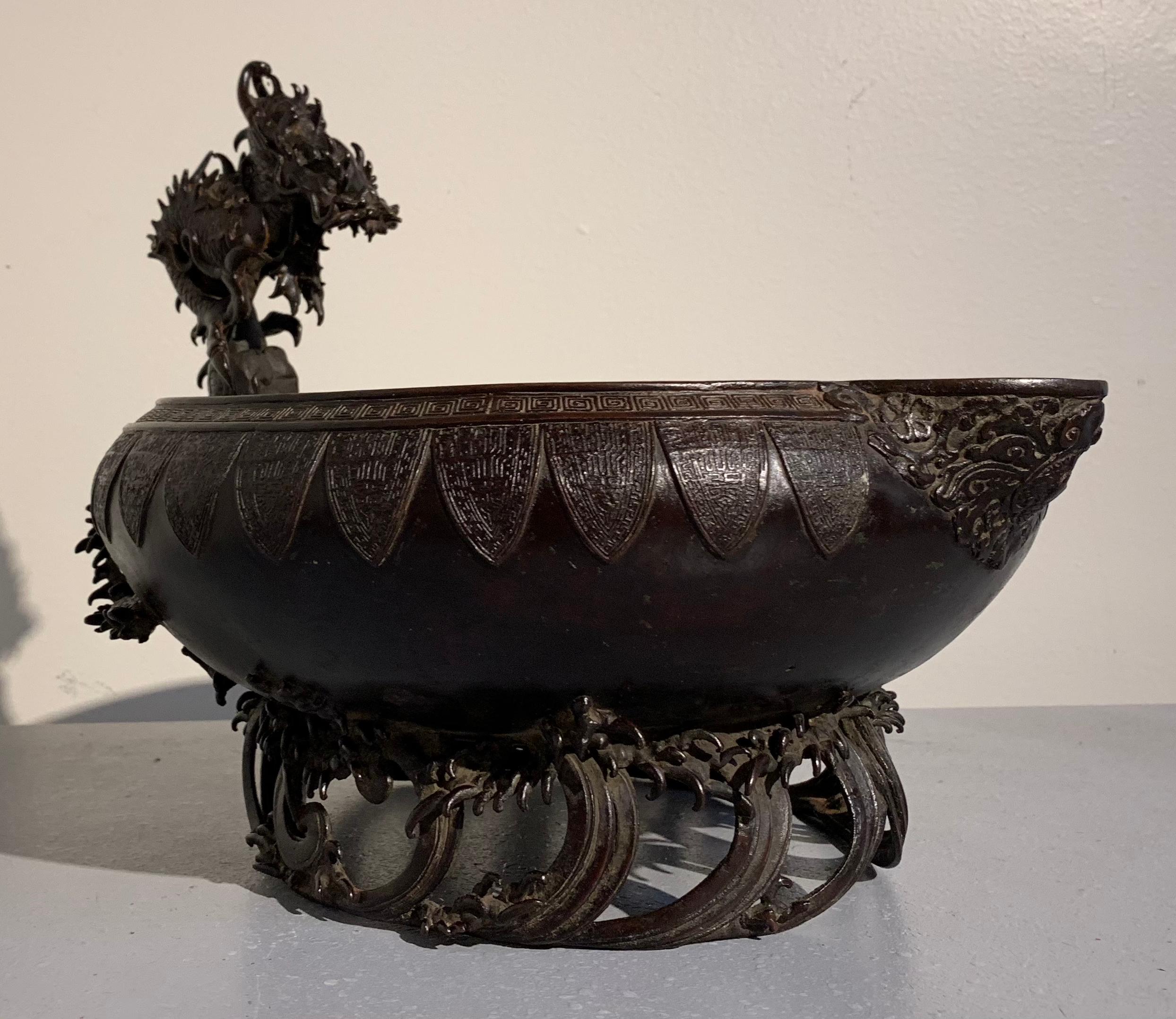 A 19th century Japanese Meiji Period bronze water basin, called a mizubachi, with archaistic designs and a powerfully cast dragon handle, all supported on a high base designed as crashing waves. 

The basin is cast with an archaistic Chinese
