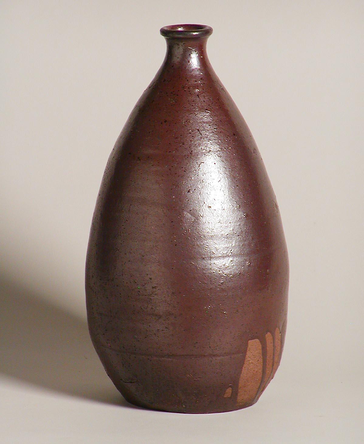 Japanese ceramic Tamba aka-dobe tokkuri, a red clay bottle vase from the Tamba kilns, a wide bodied tapering form with narrow short neck and slightly everted mouth rim covered in an iron red brown clay based glaze with a small lower section having