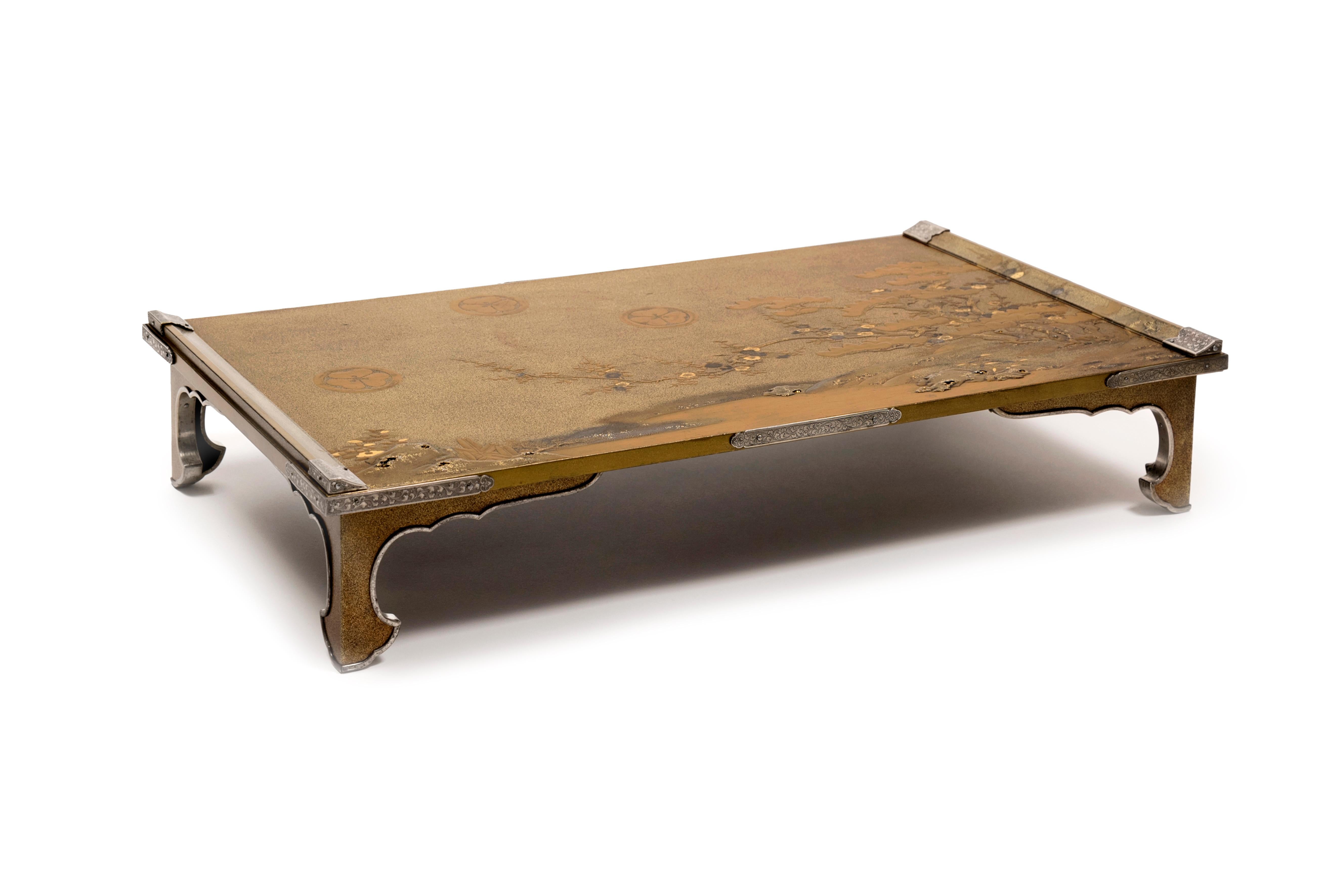 Rectangular bundai writing table on four legs in gold lacquer with black highlights on a nashi-ji background. The top is decorated with a lake landscape with plum trees, pine trees and the clover-shaped coat of arms mon (maru ni katabami) of one of