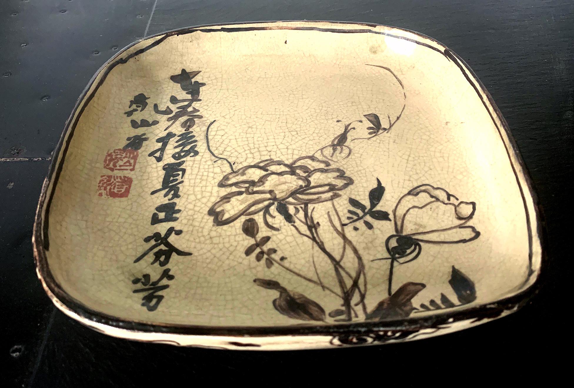 A Japanese ceramic shallow dish in square form with rounded corners from Meiji period in the style of Ogata Kenzan. The dish features a cream color crackled glaze with iron underglaze paint that evokes the aesthetic of ink painting. A bundle of