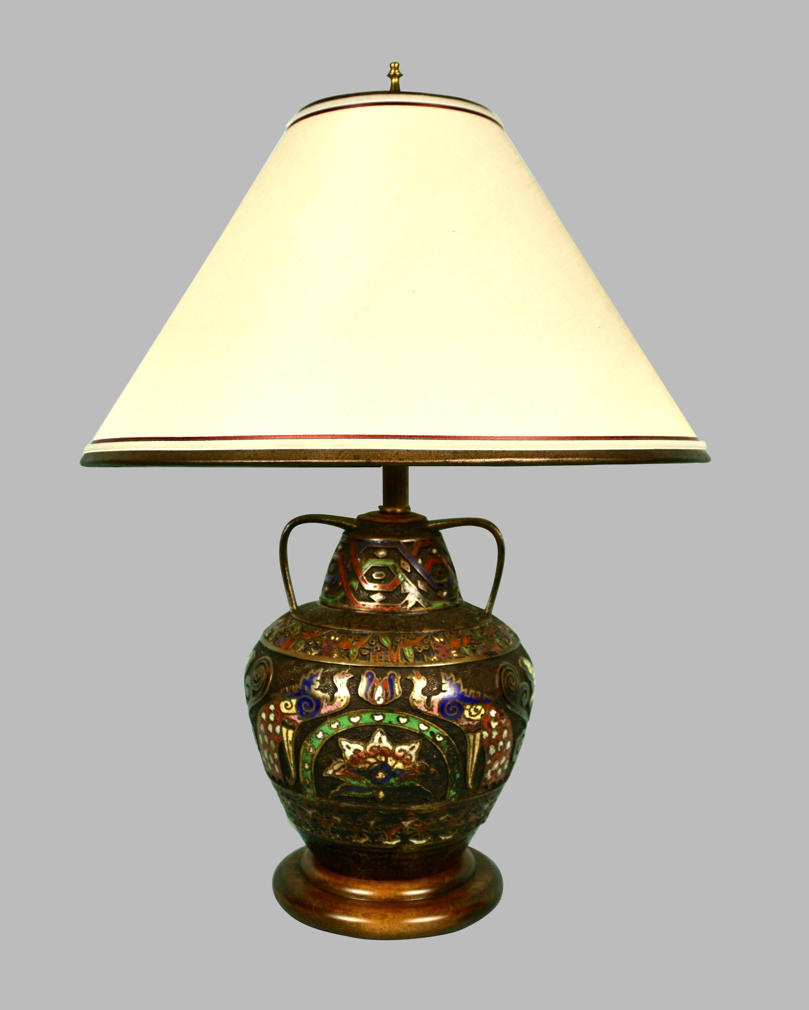 A Japanese brass champleve vase of baluster form with 2 handles decorated overall with stylized peacocks and butterflies in tones of blue and red now electrified and fitted as a lamp resting on a wooden base. Champleve is a process of filling