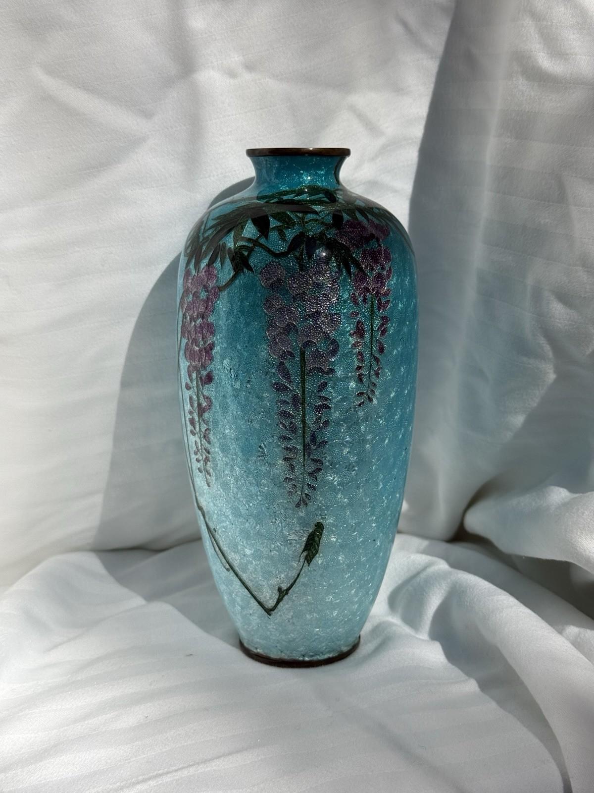 Un magnifique vase ancien en émail cloisonné japonais mesurant 7 pouces de haut. Le vase présente des fleurs de glycine en cascade dans des tons de violet et de lavande avec de délicates feuilles vertes, le tout sur un fond bleu glacier chatoyant
