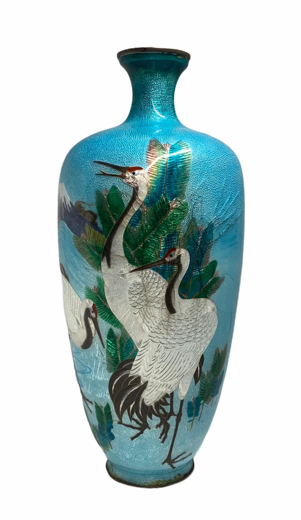 This is a cloisonné foil metal vase depicting a continuous scenes around it of three large white cranes, a volcano and some green leaves in a turquoise background. Under the base there is some Japanese mark.