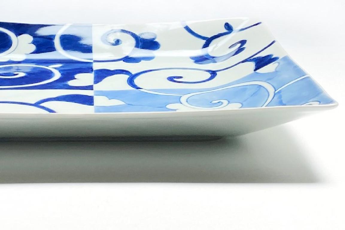 Japanese contemporary porcelain dinner plate in an elegant square shape, hand-painted in blue uderglaze in cobalt and light blue to showcase a graceful arabesque or karakusa pattern on a background of suares in blue and white. The artist's signature