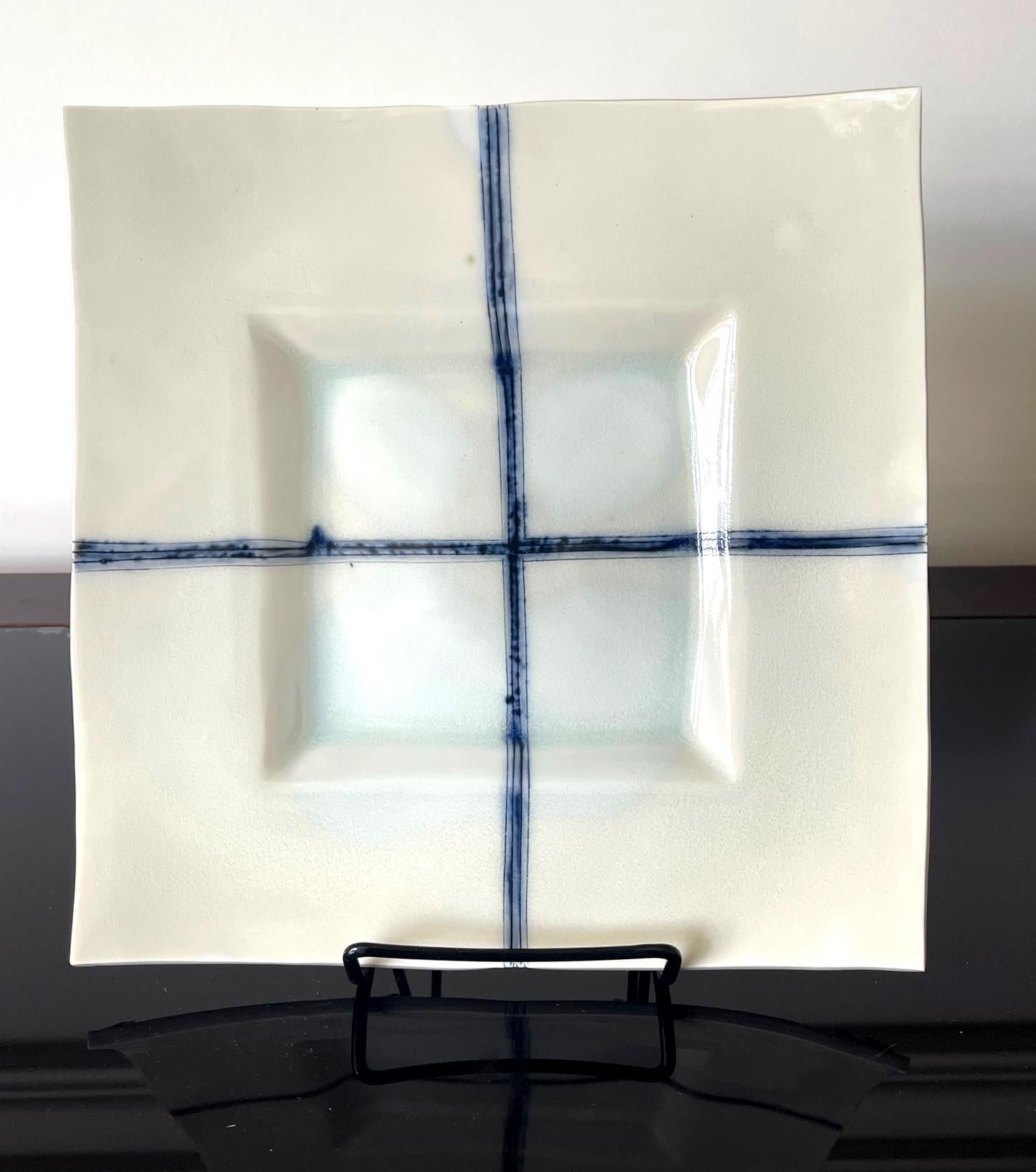 A ceramic square plate with white-celadon glaze by Japanese potter Yoshikawa Masamichi (1946-). The sharp-edged plate features a concaved center and wide border. Cobalt blue lines in bands were drawn to cross at the center. The base also has a