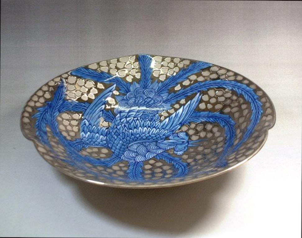 Exquisite dimpled Japanese decorative porcelain plate intricately hand painted, showcasing a dramatic breathtaking phoenix in deep blue extending its beautiful long wings to cover the entire body of the charger, set against a stunning platinum