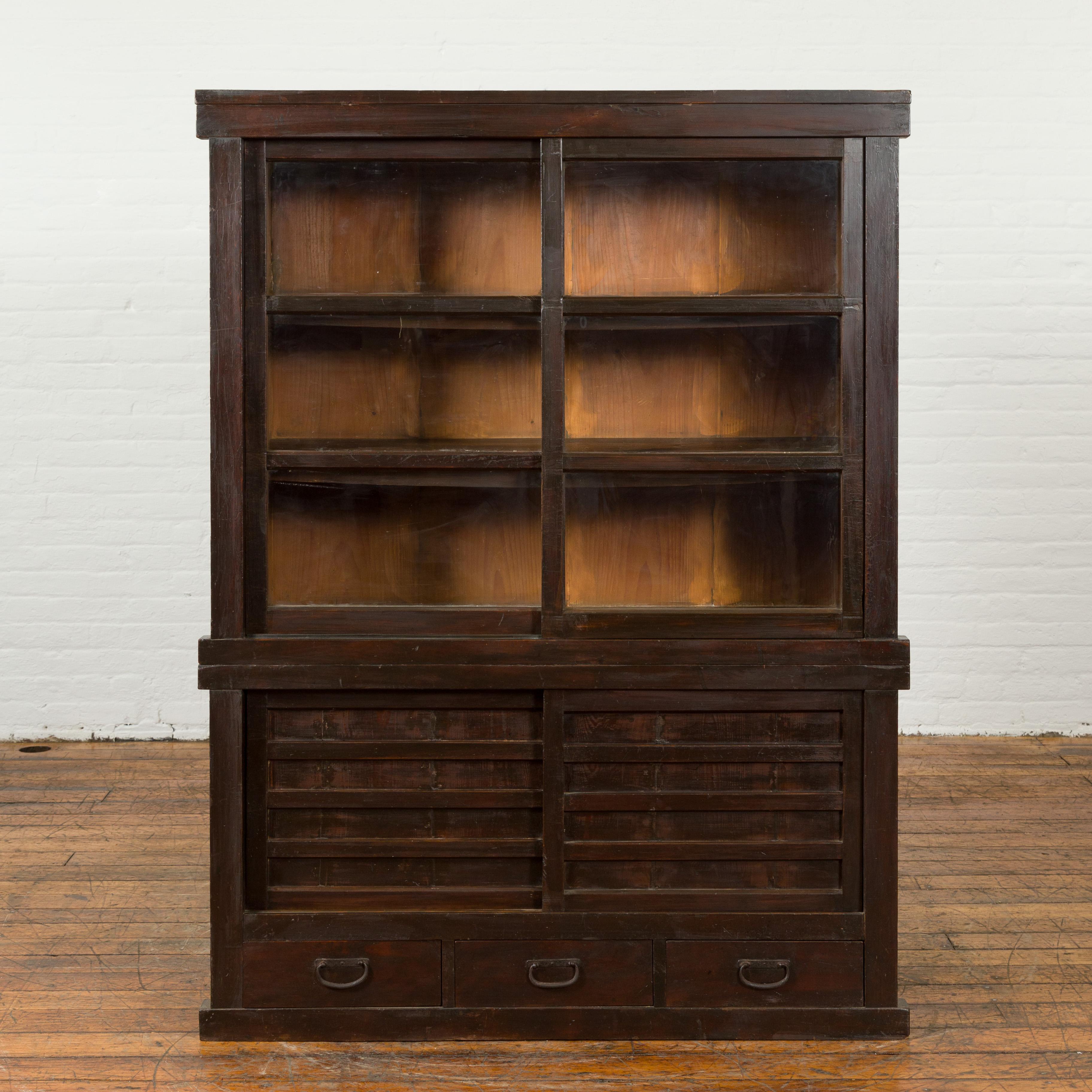 A Japanese wooden bookcase from the early 20th century with glass sliding doors and drawers. Created In Japan during the second quarter of the 20th century, this wooden bookcase features a linear Silhouette perfectly complimented by a dark brown