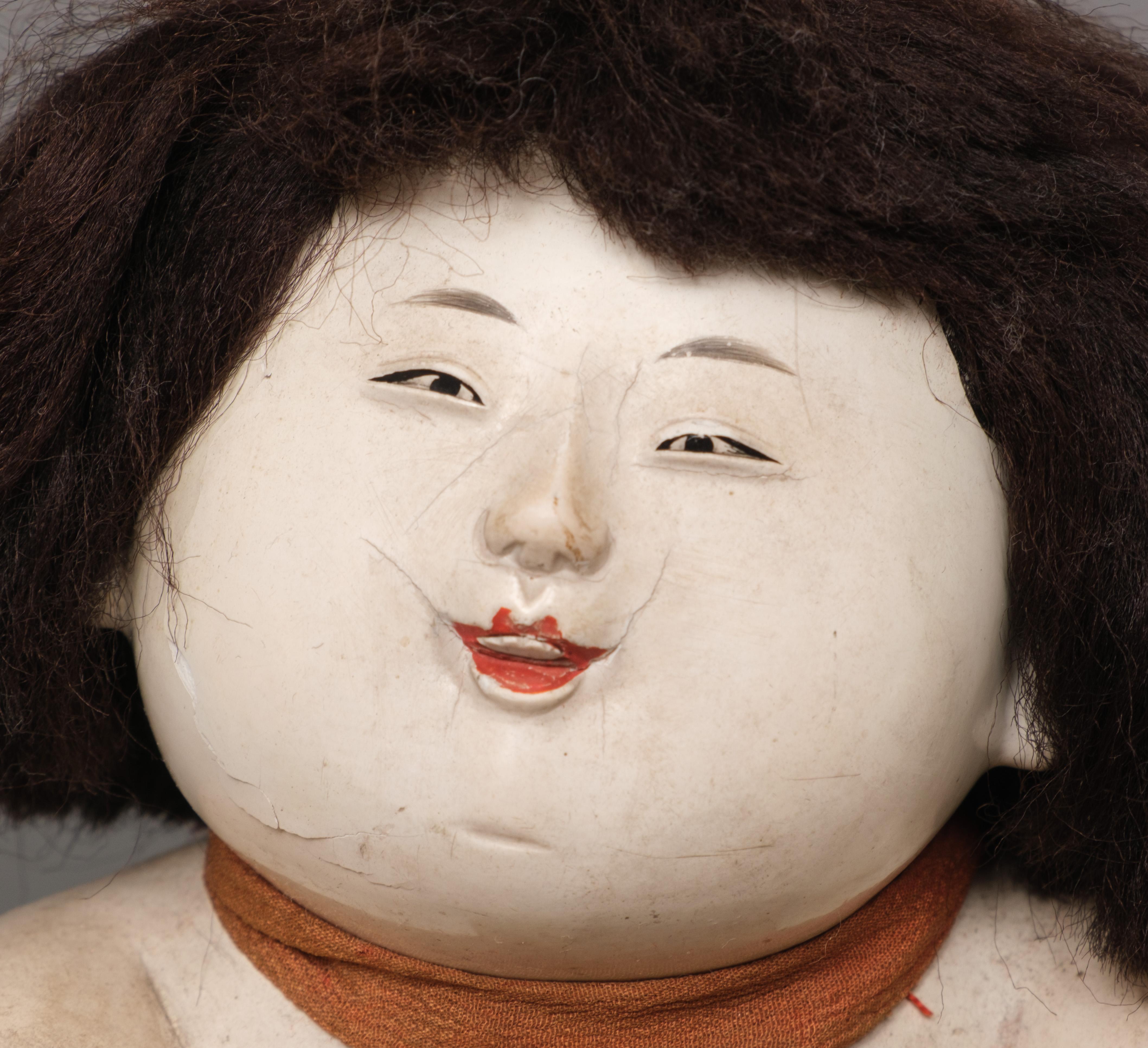 A charming gosho’ningyô (palace doll) of plump, seated child with a brilliant white skin and a small delicately elegant face, reminiscent of a young noble. The doll is playfully raising its left hand to display a golden ball, pointing at it with a