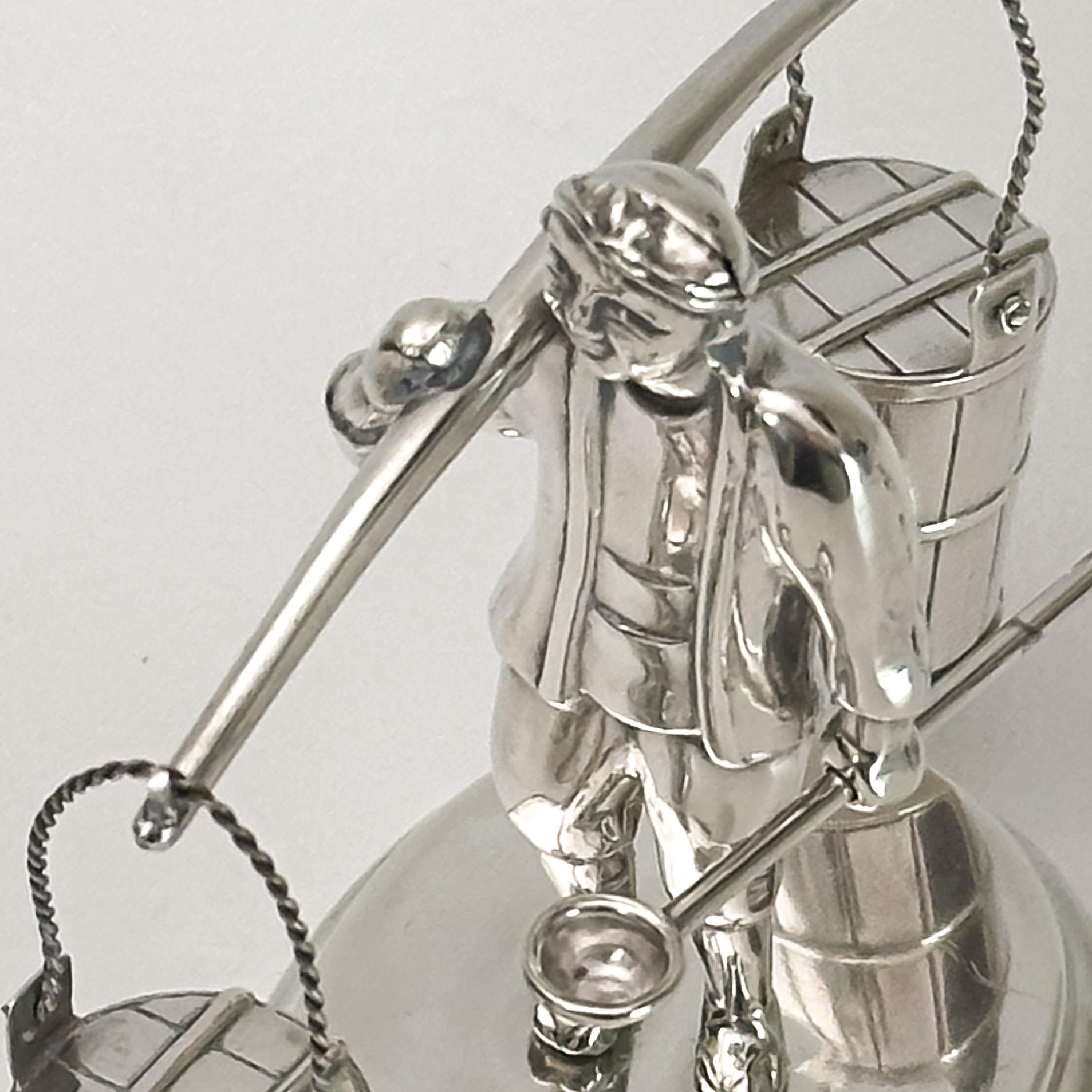 A whimsical salt and pepper set crafted in Japan during the 1950s, made entirely of sterling silver. 

The set features a charming figure of a Japanese man standing atop a decorative oval-shaped plinth. He holds a long shoulder yoke balanced with