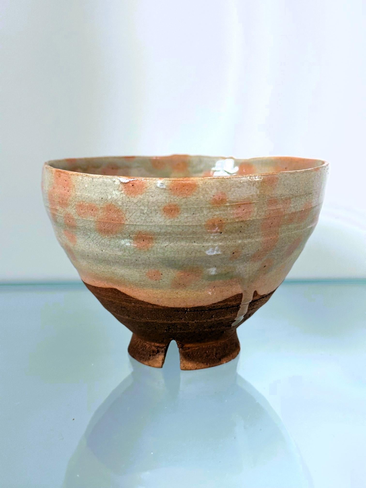 A bespoken Japanese ceramic tea bowl with glazed with fabric insert, pouch and original padded wood tomobako box. The chawan has a slightly irregular wall supported by a high notched foot ring. The glaze features a distinct pinkish spotty pattern on