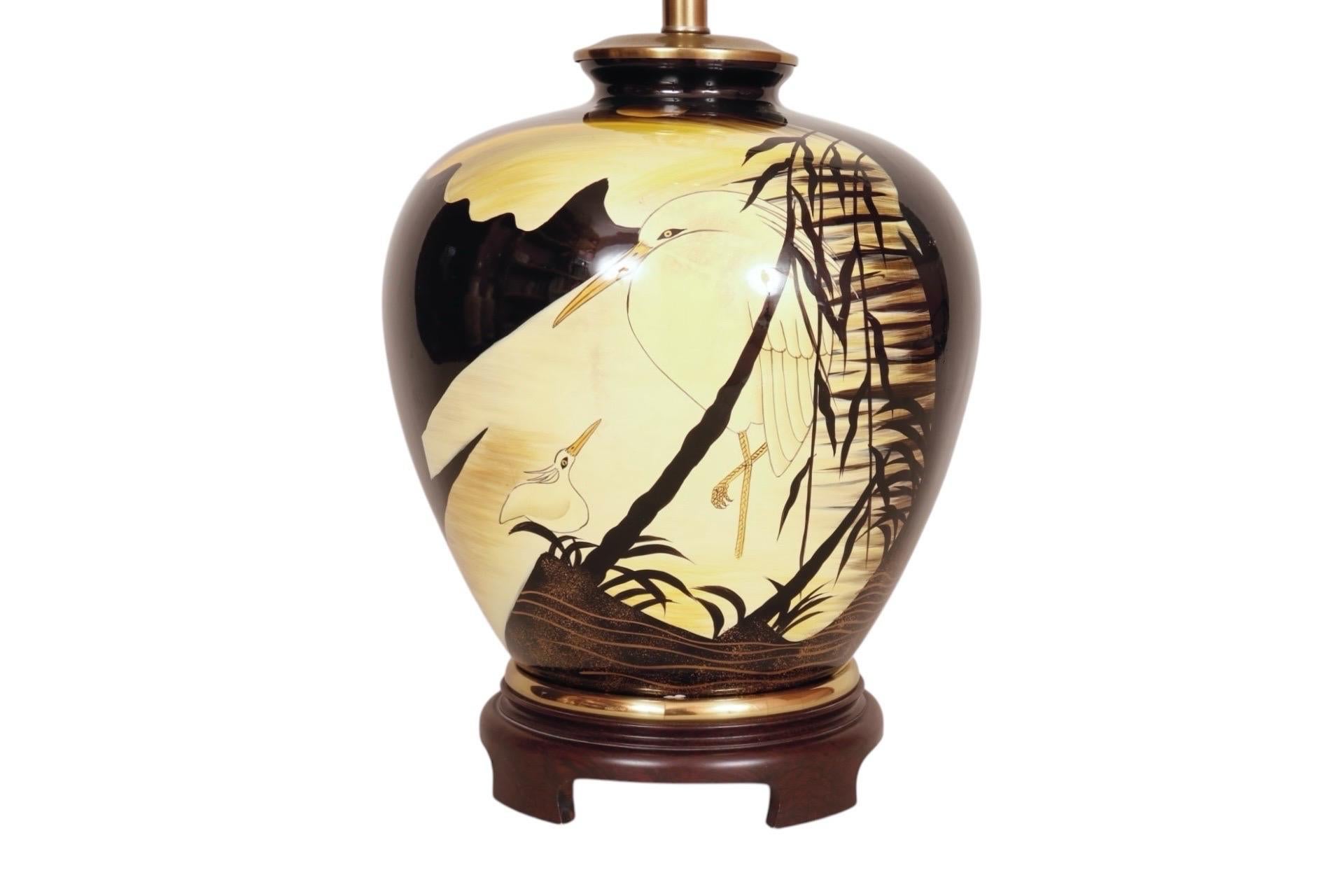 Una lampada da tavolo in ceramica giapponese dipinta a mano. Il vaso è decorato su entrambi i lati con una madre e un piccolo airone nell'acqua, con steli di bambù in silhouette. L'arpa è sormontata da un'urna e sotto la presa c'è un cappuccio in