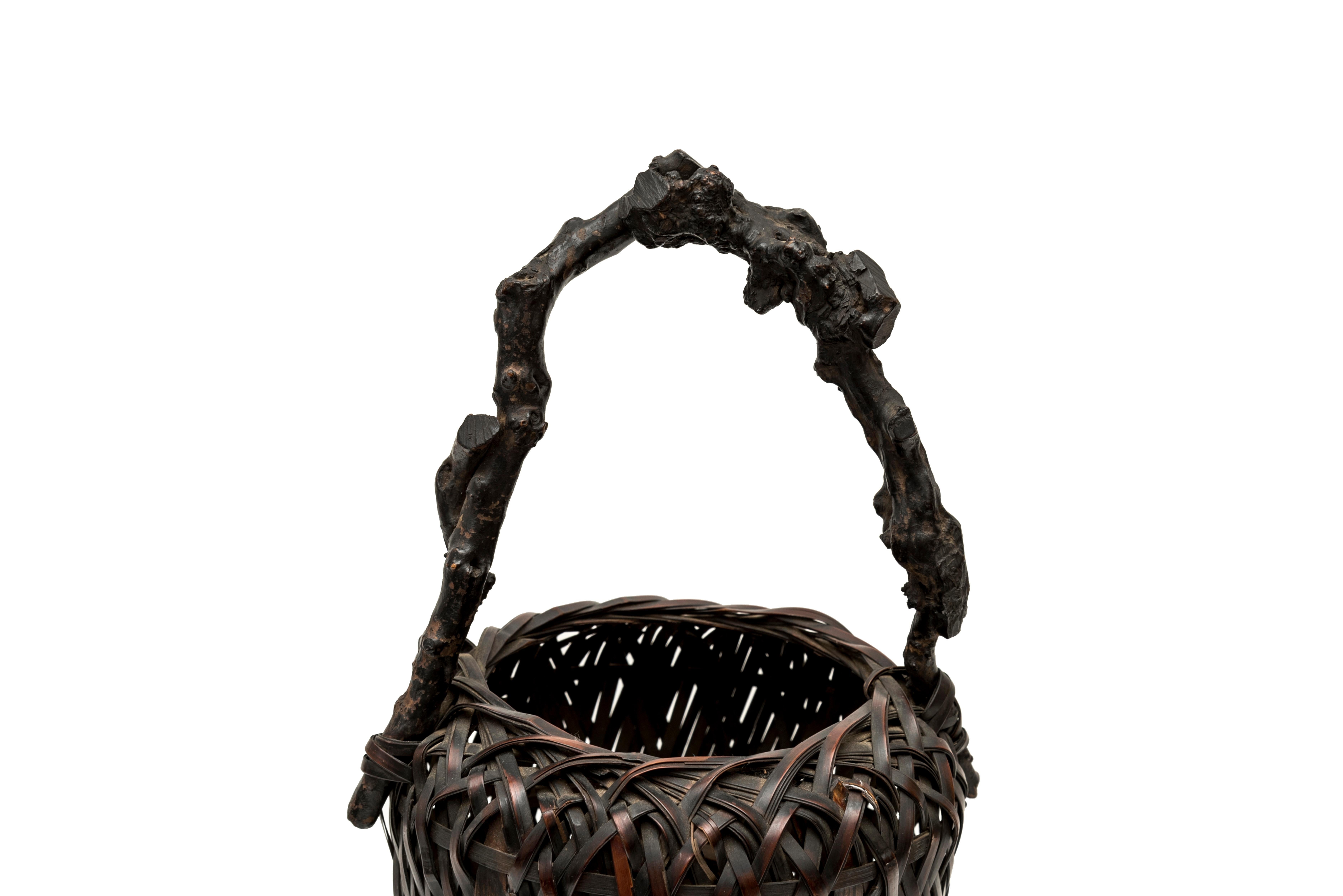 Ikebana basket (hanakago) made of dark brow varnished bamboo wickerwork, elongated rounded shape, with a handle made of three varnished wooden branches. Thicker bamboo braids on the four sides. The roughness of the handle is a reminder of a raw wood